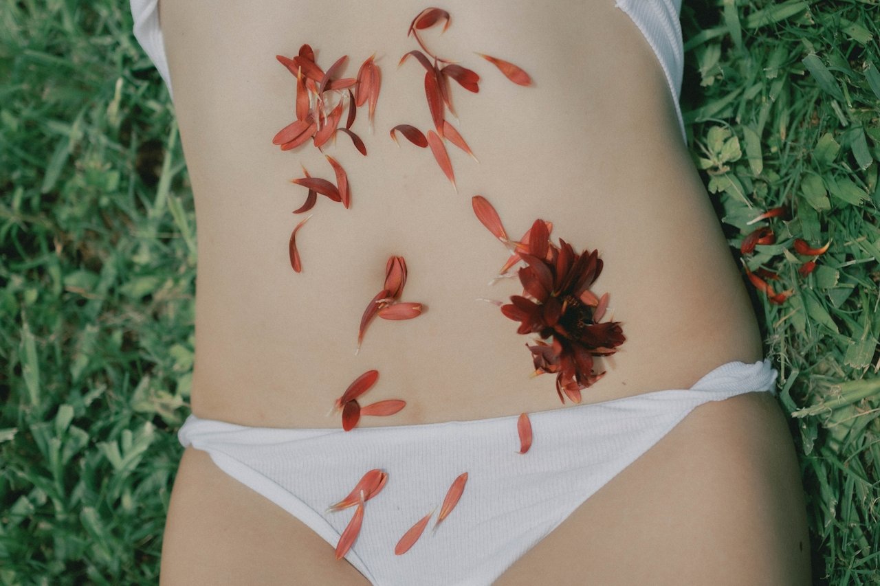 A person lying on grass, wearing white underwear, with red flower petals and a red flower arranged on their stomach and hip area.