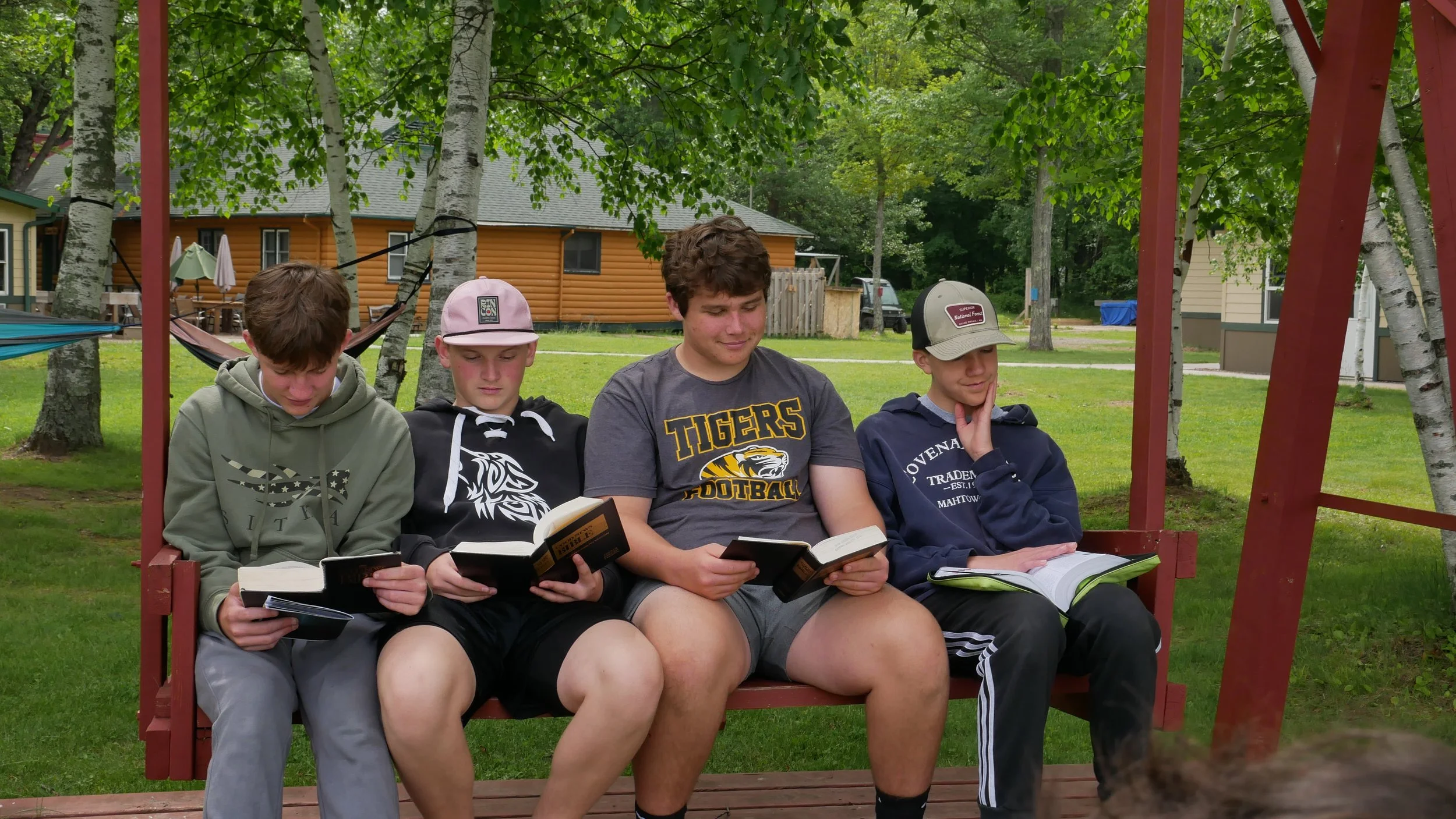 Four teenage boys sitting on a red wooden swing with books in their hands, in a grassy backyard with trees and small houses.