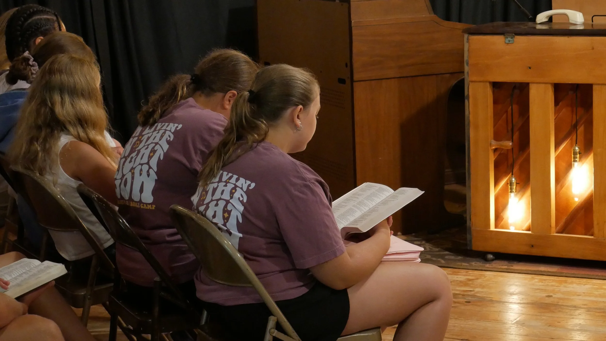 A group of young girls sitting in chairs, reading or praying with hymnbooks or Bibles, in a rustic indoor setting with wooden floors and warm hanging lights.
