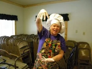 Young girl dressed as a chef, wearing a white chef's hat and apron, holding a large spoon and smiling.