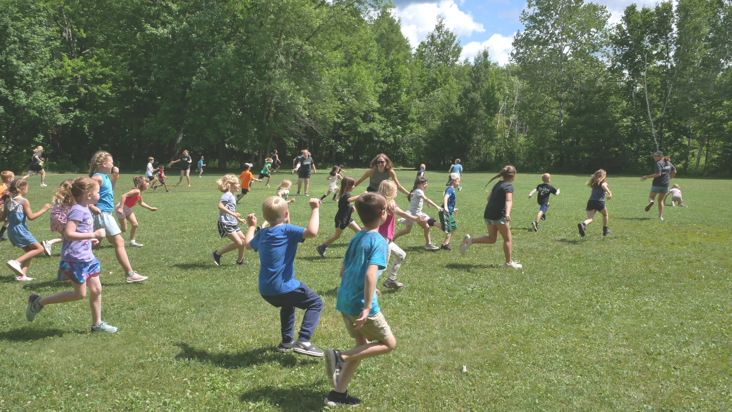 Group of children and adults running and playing on a grassy field in a park during sunny weather.