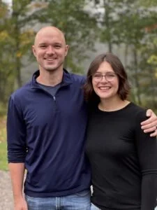 A smiling man and woman standing outdoors with trees in the background, embracing each other.