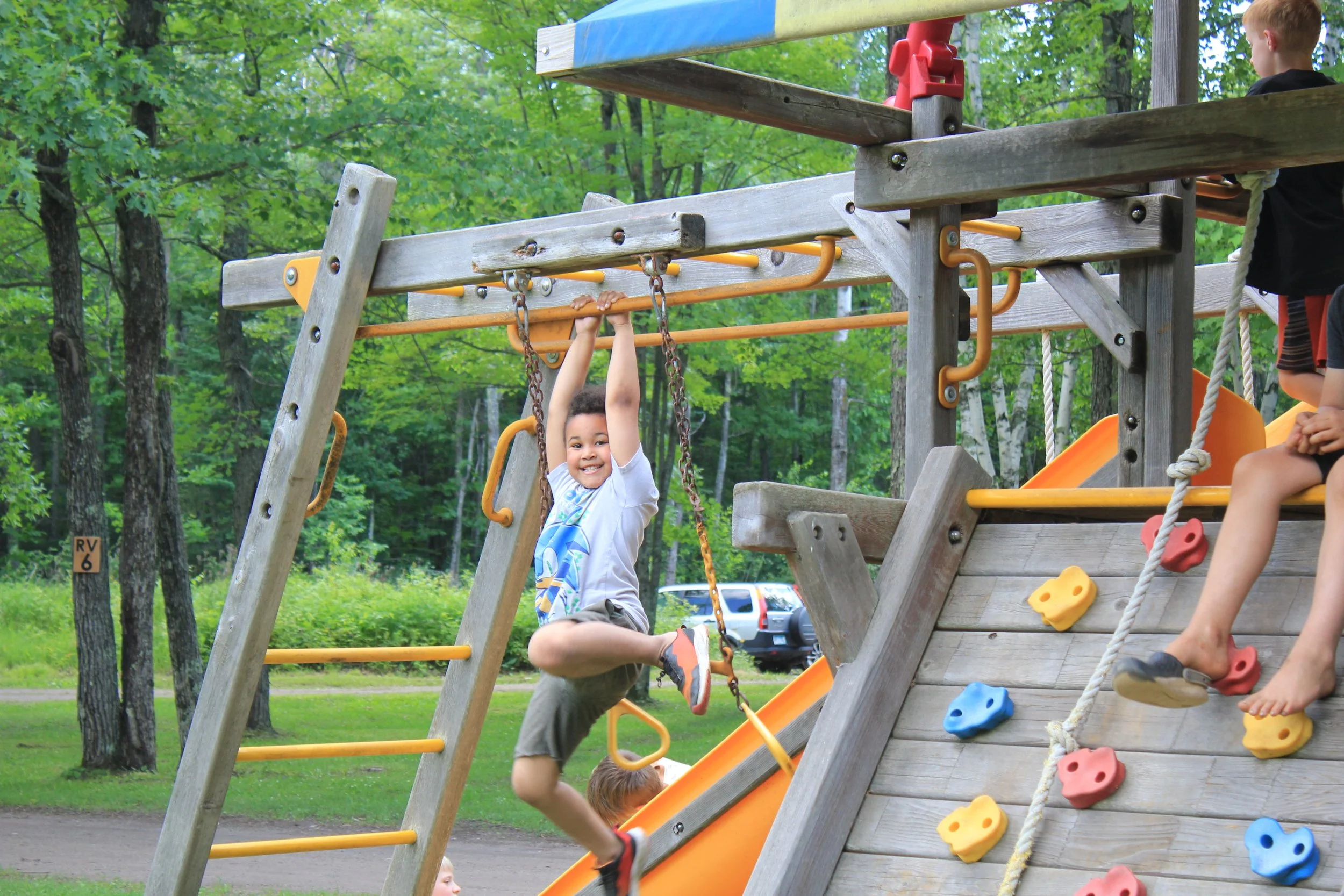 Children playing on a wooden playground structure in a park with trees in the background. One boy is hanging from a bar, smiling at the camera, while another child is sitting nearby on the structure.