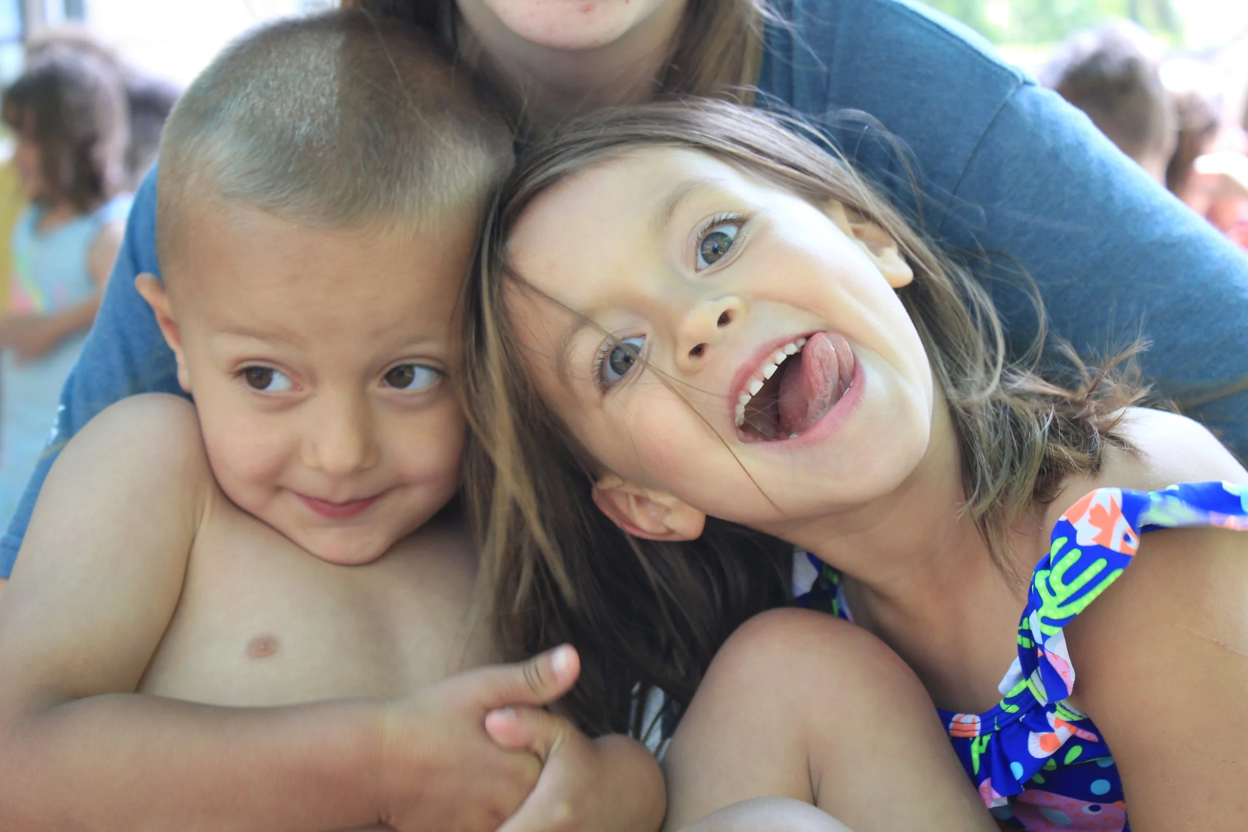 Two children and an adult showing playful expressions; one child smiling softly, the other sticking out tongue and making a funny face, outdoors with other kids in the background.
