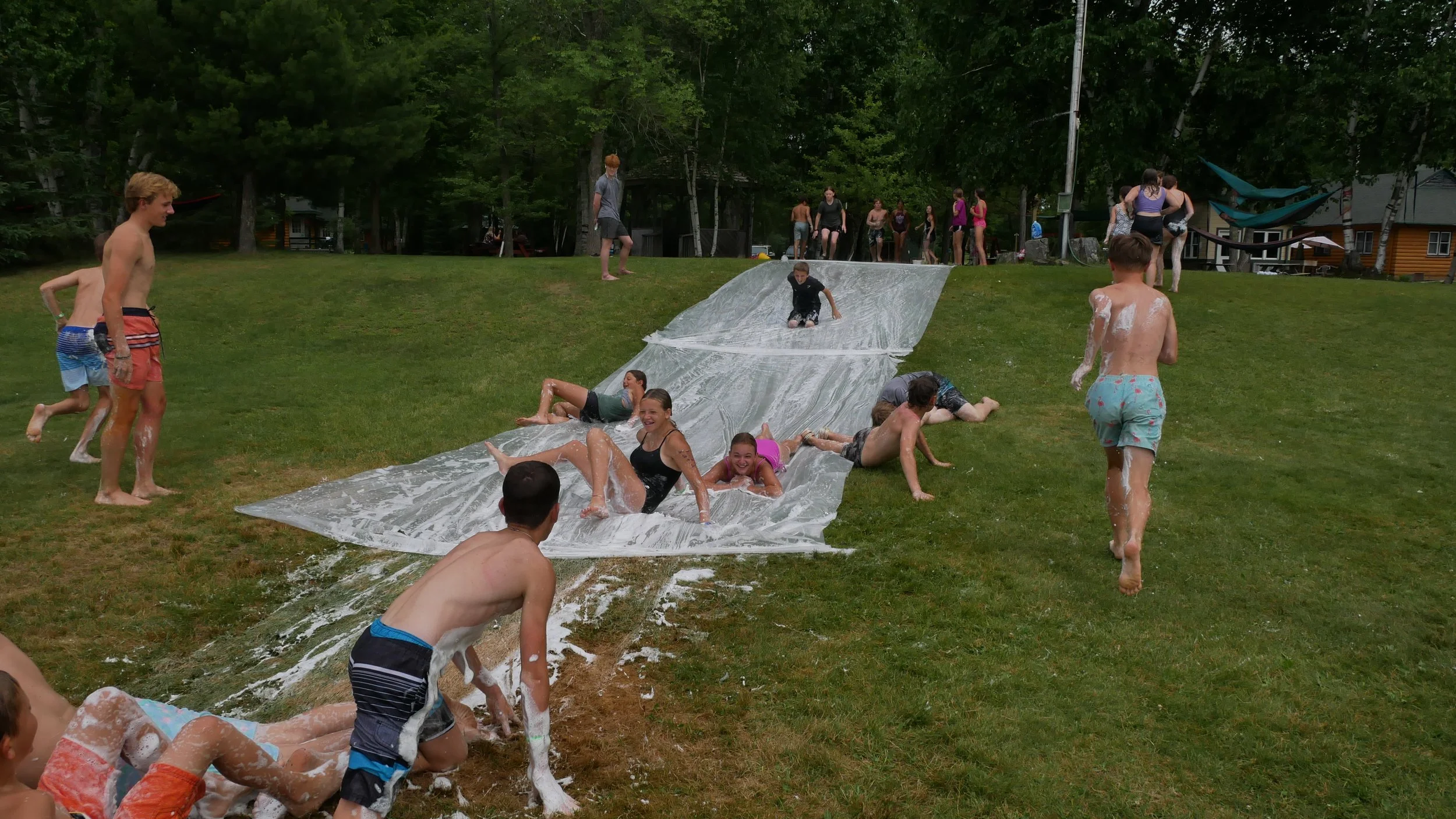 Children and teenagers playing on a slip 'n slide on a grassy yard, with some sliding and others running towards it, surrounded by trees and adults in the background.