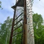 A wooden outdoor climbing structure with a climbing wall and a rope swing, surrounded by trees and greenery.