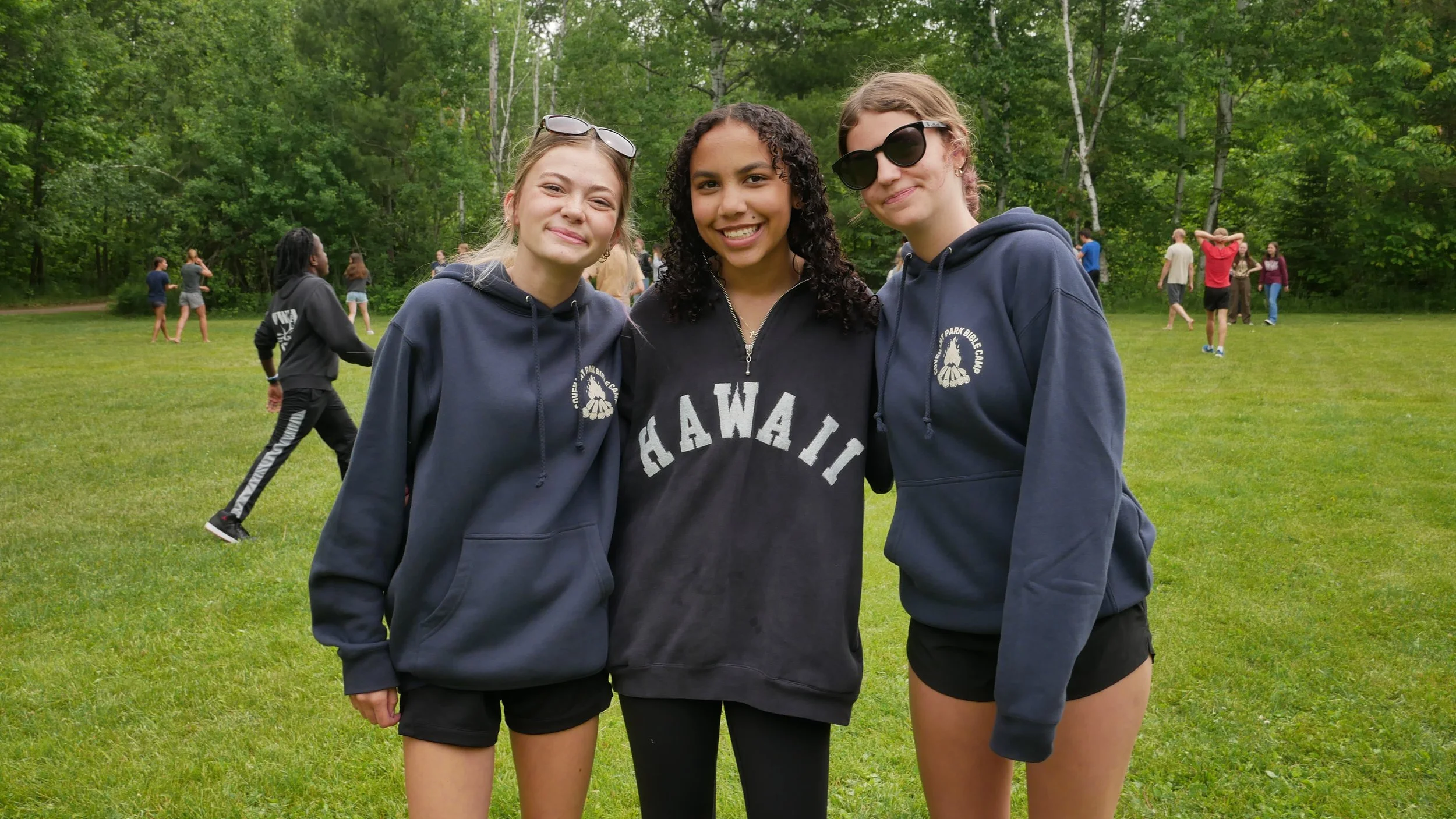 Three teenage girls stand together outdoors on a grassy field, smiling at the camera. They are wearing hoodies, with the middle girl wearing a black Hawaii sweatshirt and the other two in navy blue hoodies. In the background, other children are playing and walking around among trees.