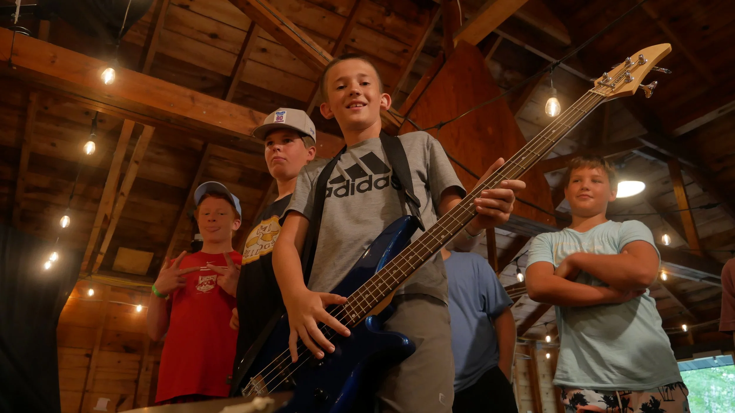 Group of boys at an indoor gathering. One boy in the center is holding a blue electric bass guitar, smiling at the camera. The other boys around him are making various facial expressions and poses. The setting has a wooden ceiling with string lights.