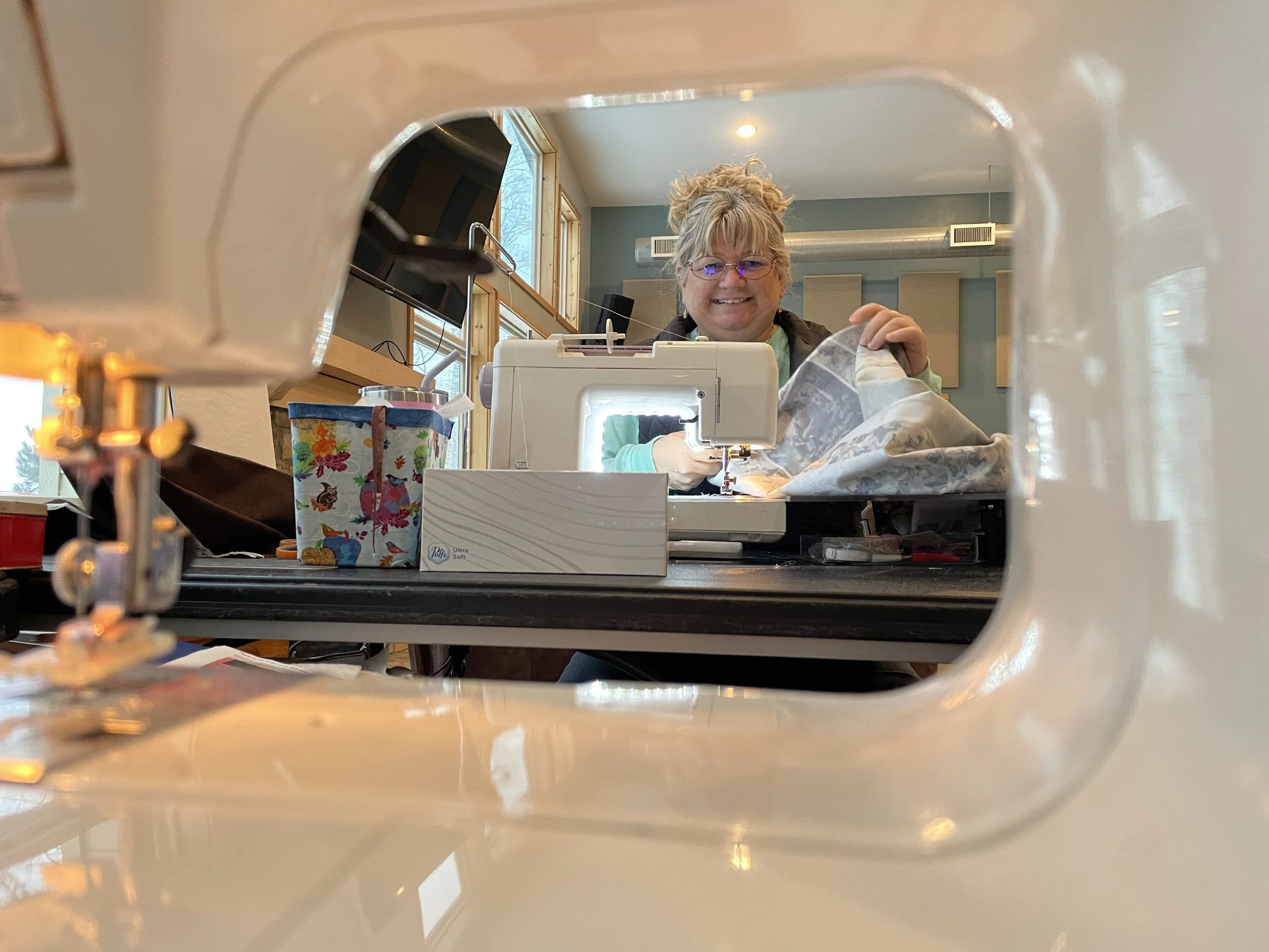 Woman smiling and working with a sewing machine, visible through the fabric arm of the sewing machine from underneath, with colorful sewing supplies on the table and a cozy, well-lit room in the background.