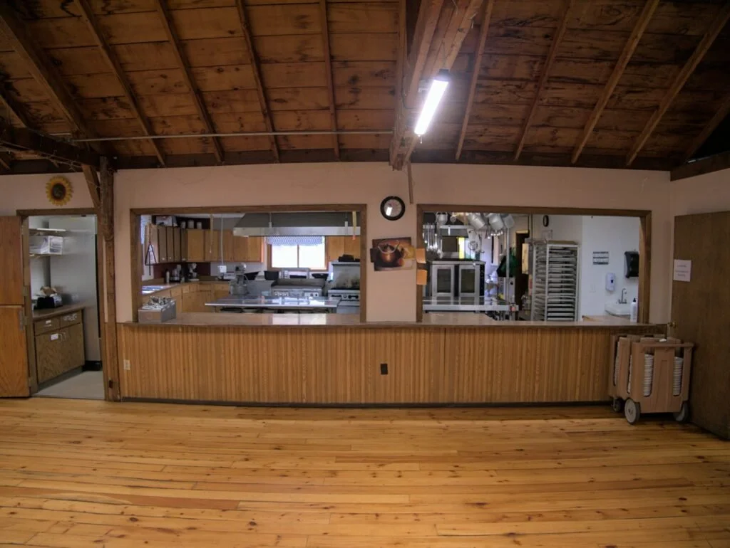 View of a rustic kitchen seen through an open serving window, with wooden walls and ceiling, stainless steel appliances, and a wooden floor.