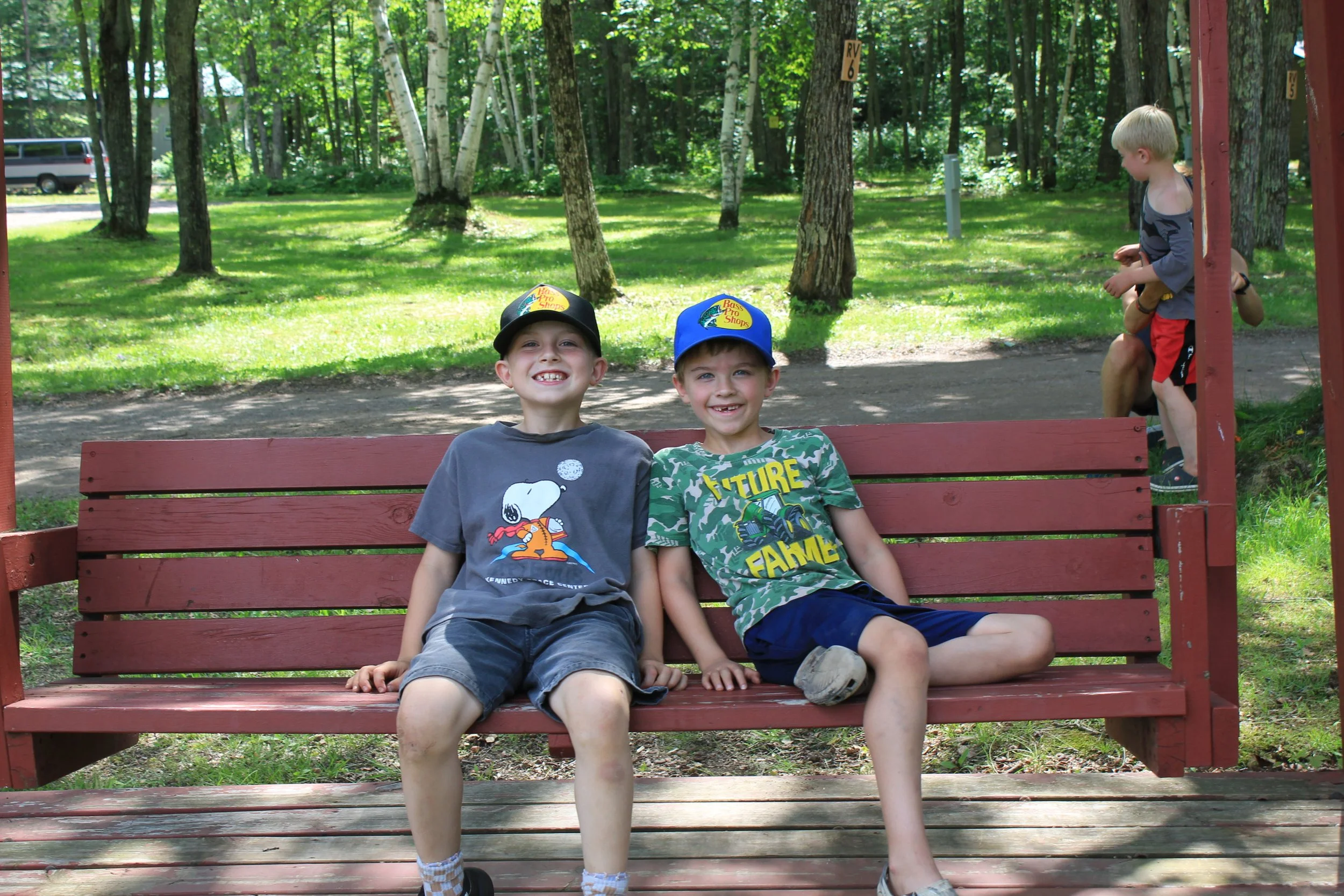 Two young boys sitting on a red park bench in a wooded area smiling at the camera, with one boy in a gray t-shirt and the other in a green camouflage shirt. Two other children are in the background, one sitting and the other standing, near a slide.