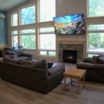 Living room with large windows, two gray sofas, a wall-mounted TV showing a scenic landscape, a bookshelf, and a fireplace.