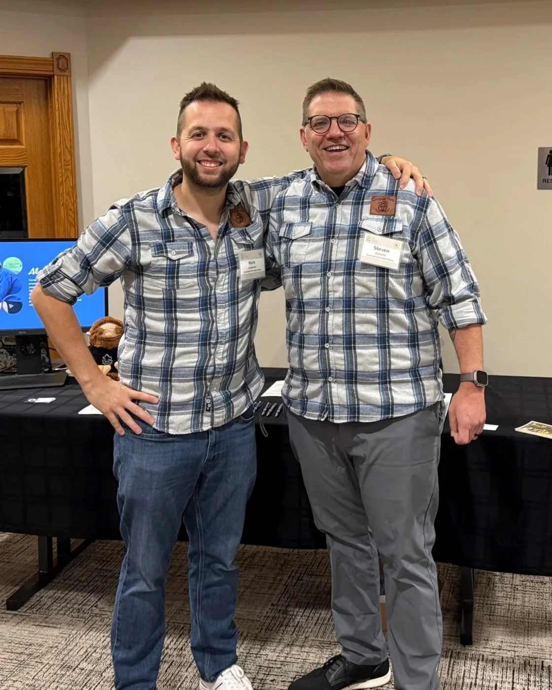 Two men in plaid shirts smiling, standing with arms around each other in a room with a black table and a monitor in the background.