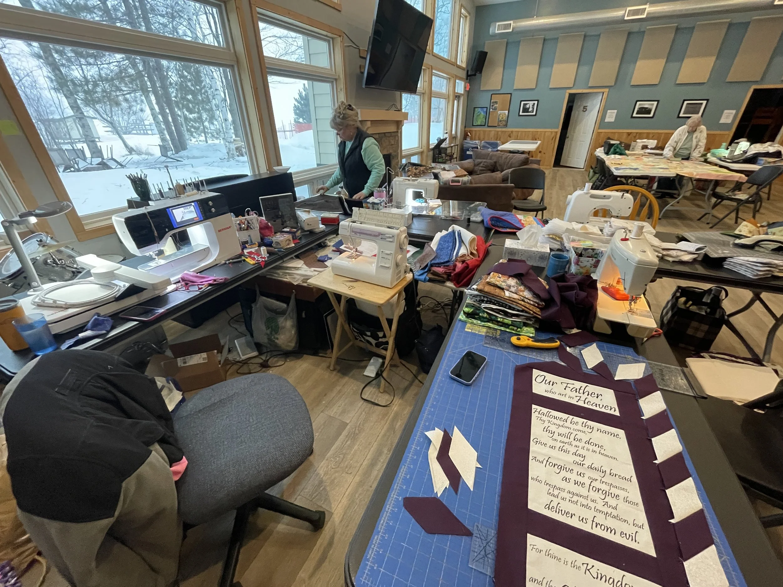A sewing and craft workspace with tables filled with fabric, sewing machines, and tools, inside a room with large windows overlooking a snowy outdoor landscape.