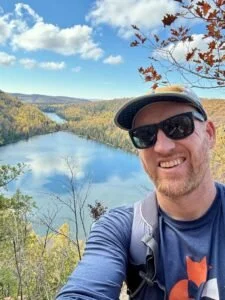 Man wearing sunglasses and a hat hiking outdoors, with a river and trees in autumn foliage in the background.