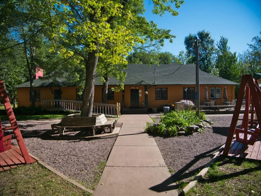 A yellow house with a dark roof, surrounded by trees, a pathway leading to the front door, outdoor benches, and a wooden swing set in the yard on a sunny day.