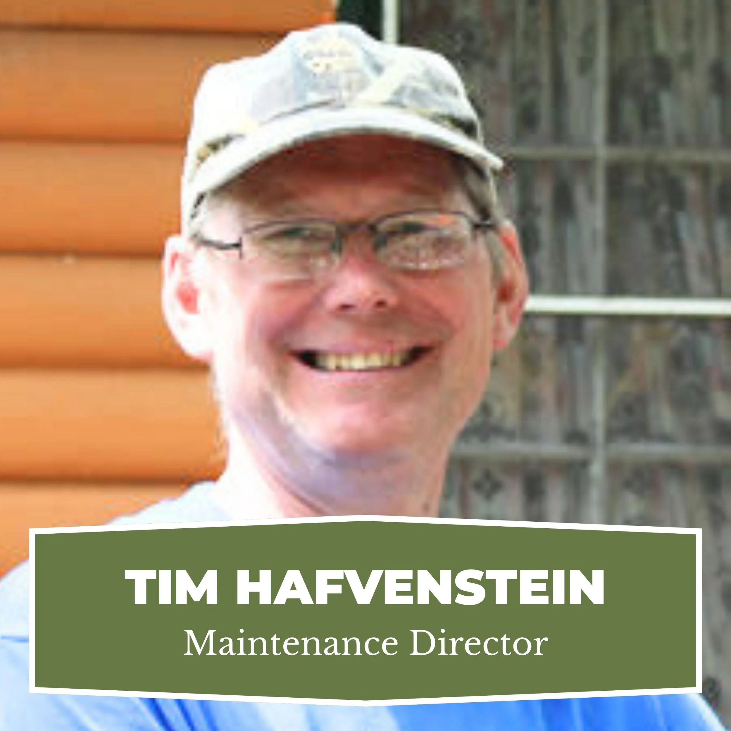 Close-up of a smiling man wearing glasses and a camouflage cap, with a wooden house and trees in the background.