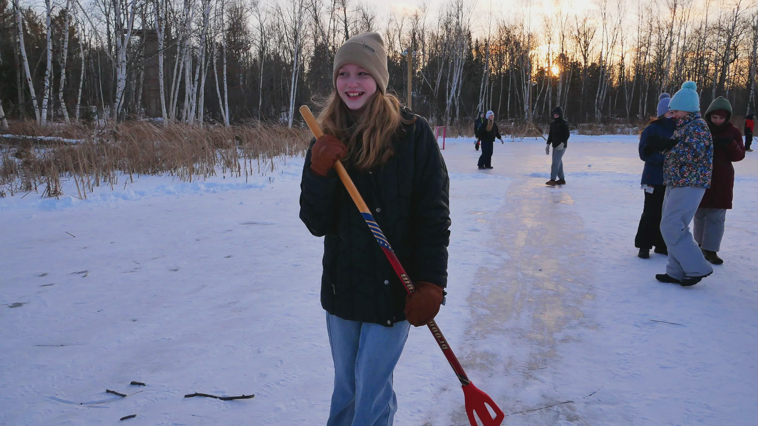 A girl with long hair wearing a beige beanie, black jacket, and brown gloves holds a red hockey stick on a snow-covered ice rink during sunset, with several people in winter clothing skating in the background near leafless trees.