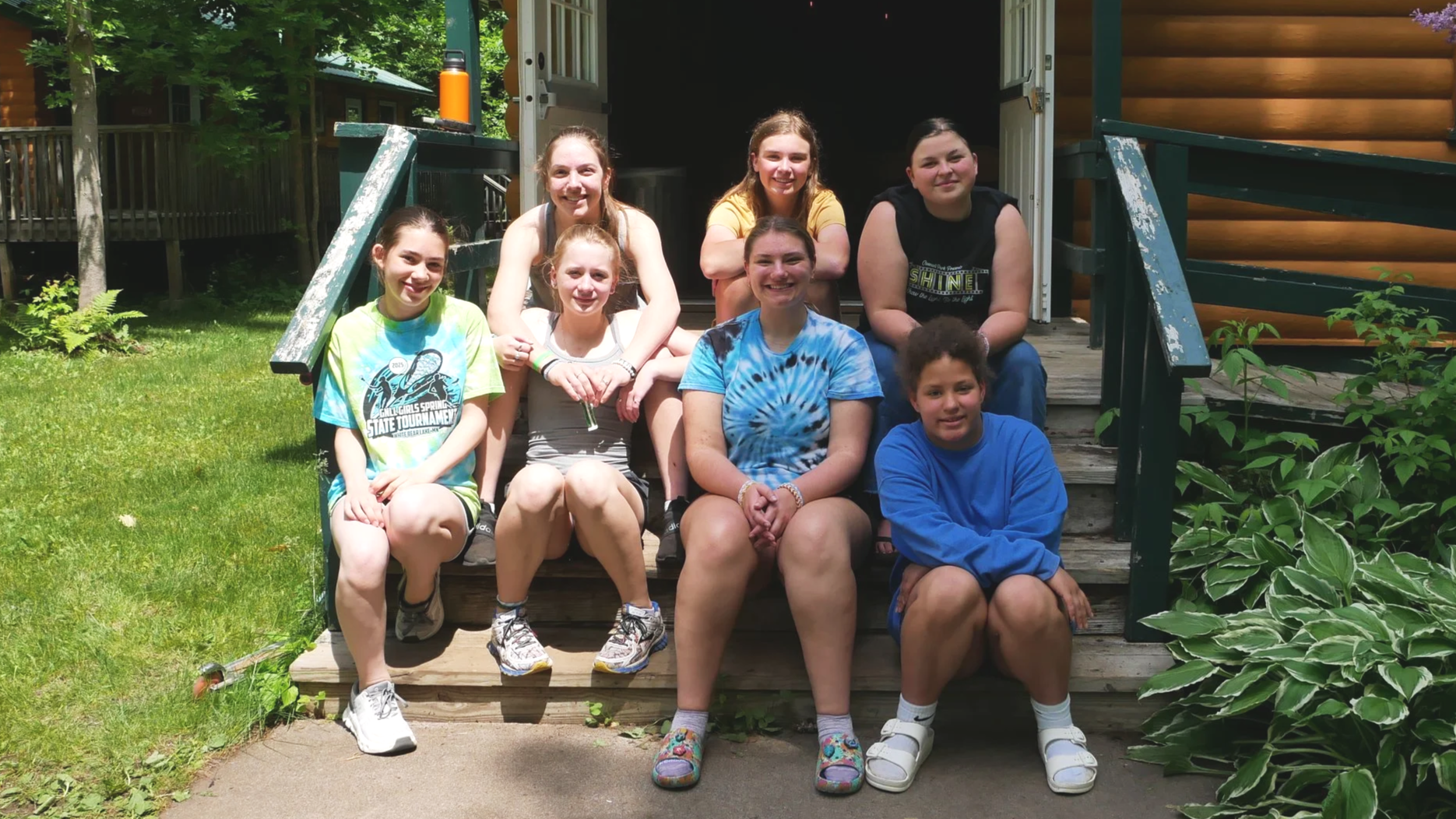 Group of nine young girls sitting on wooden stairs outside a log cabin, smiling at the camera.