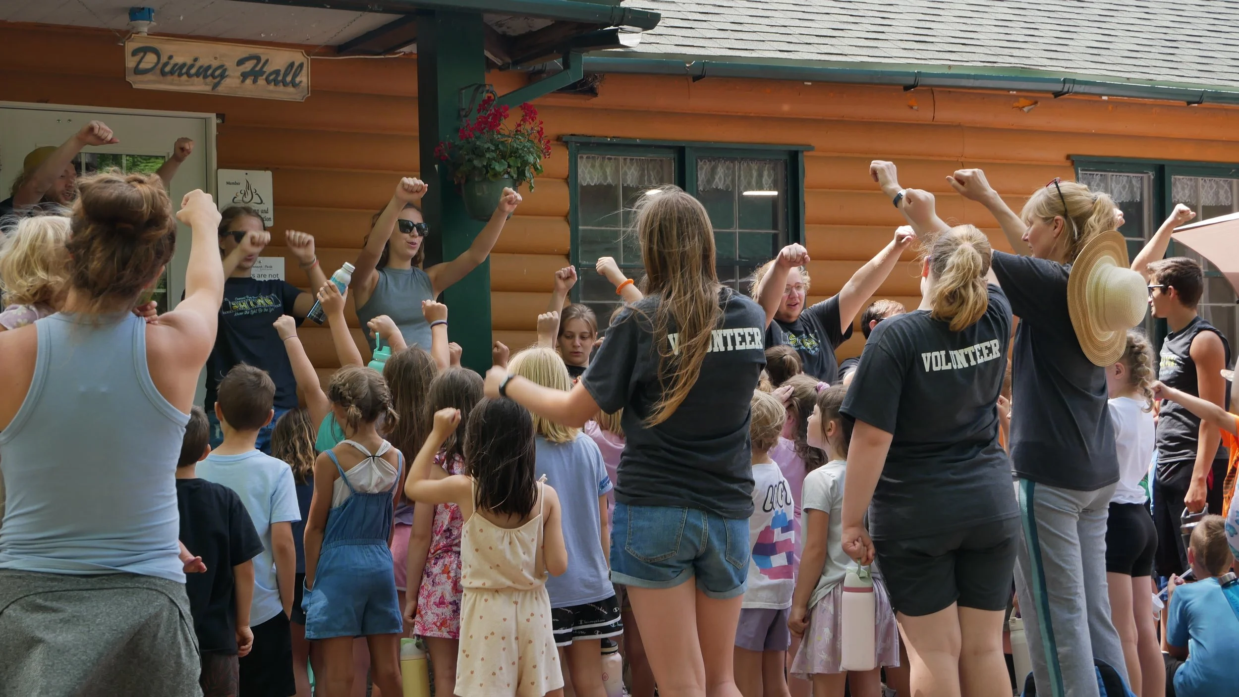 Group of children and volunteer staff at an outdoor event, raising their fists in celebration or encouragement, in front of a log cabin-style building with a 'Dining Hall' sign.