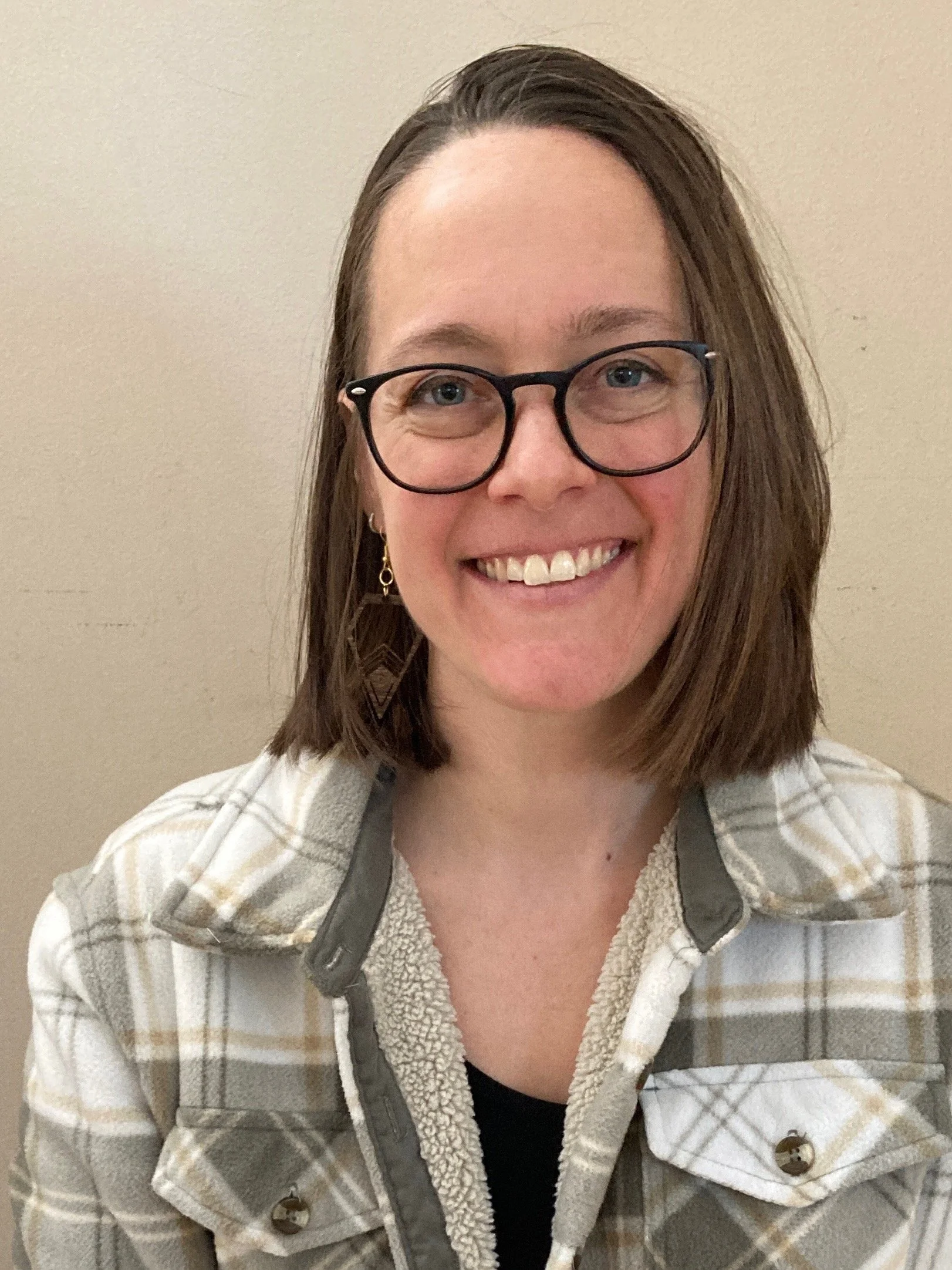 Portrait of a woman with shoulder-length brown hair, wearing glasses, earrings, and a plaid jacket, smiling against a beige background.