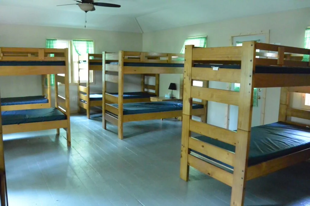 Room with multiple wooden bunk beds and small windows with green curtains.
