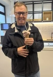 Man in glasses and black jacket holding a drink with whipped cream in a cafe or bakery.