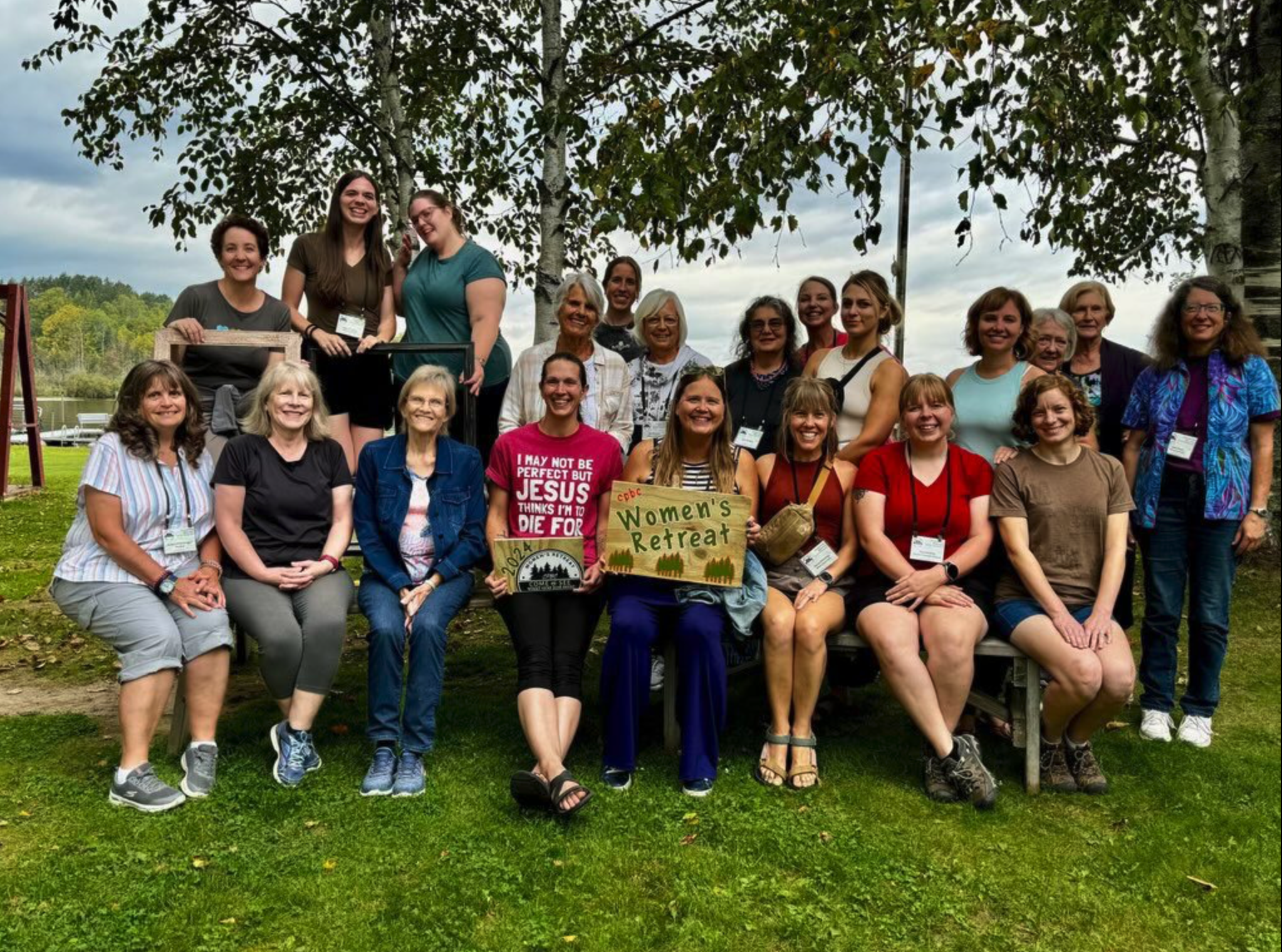 Group of women and girls gathering outdoors for Women's Retreat, standing and sitting on grass with trees and lake in background, some holding signs and wearing name tags.