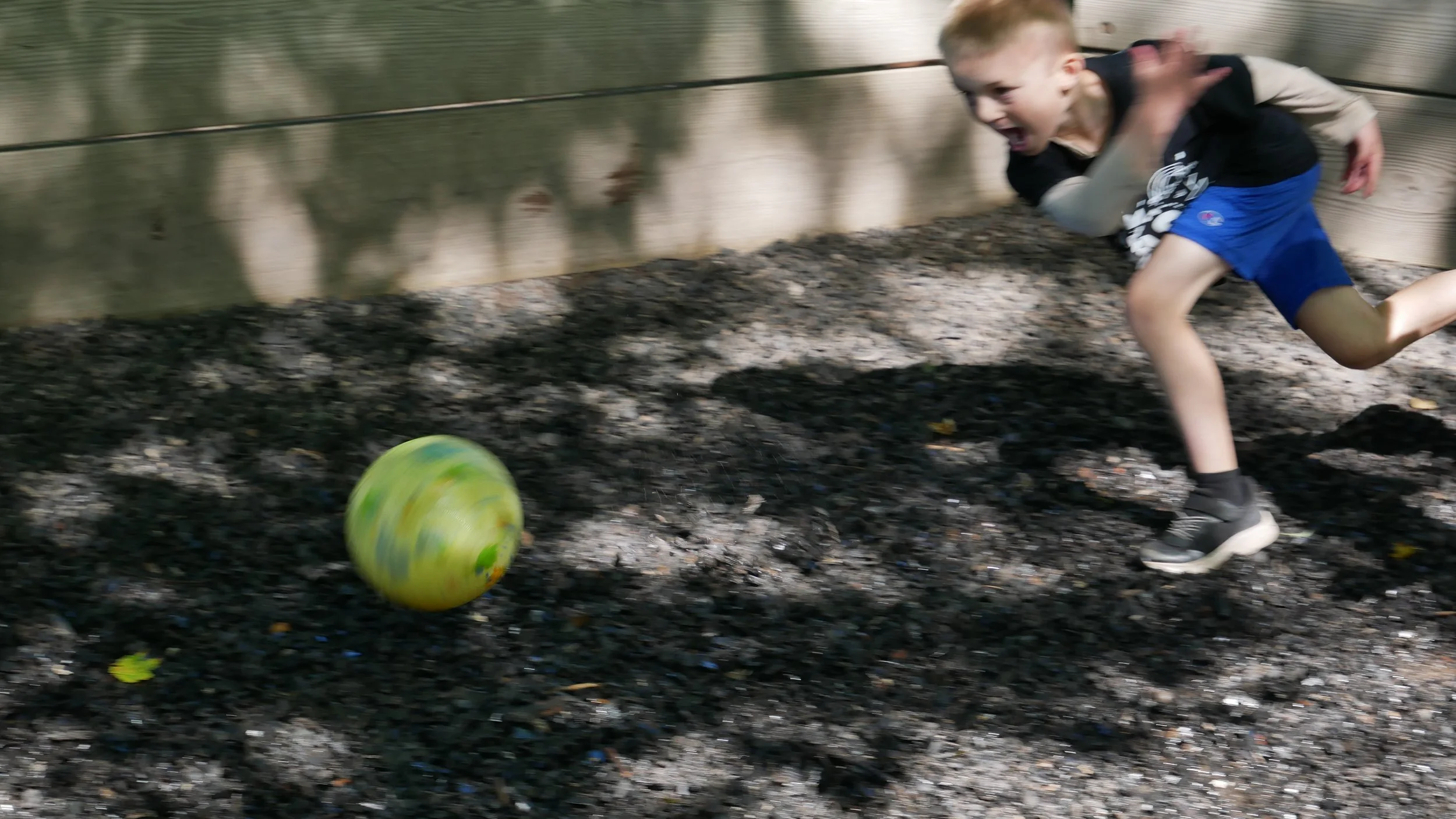 A young boy running outdoors on dark gravel, chasing a yellow and green soccer ball, with a wooden fence in the background.