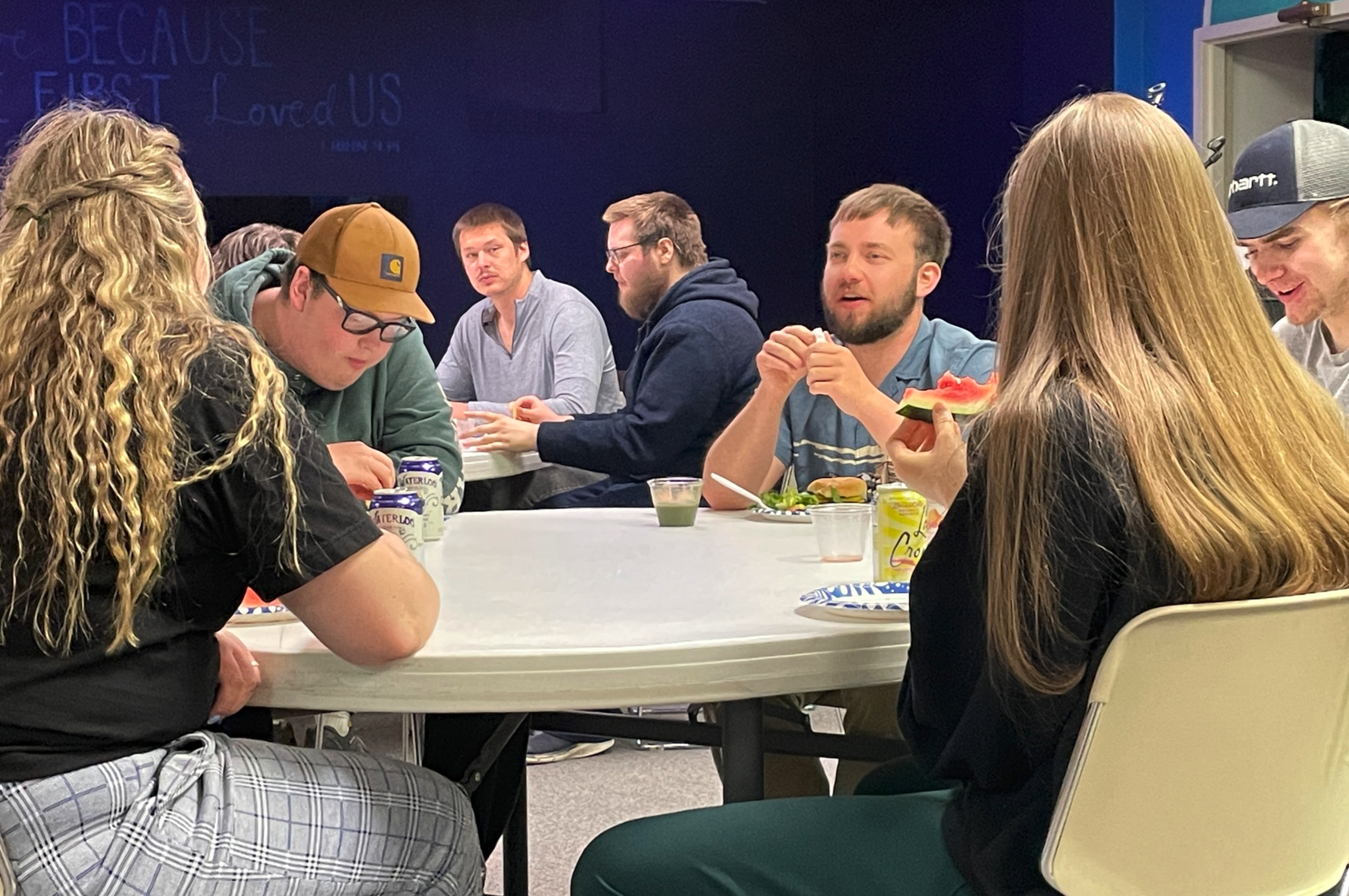 Group of young adults sitting around a white table, eating and talking, with a blue wall in the background.