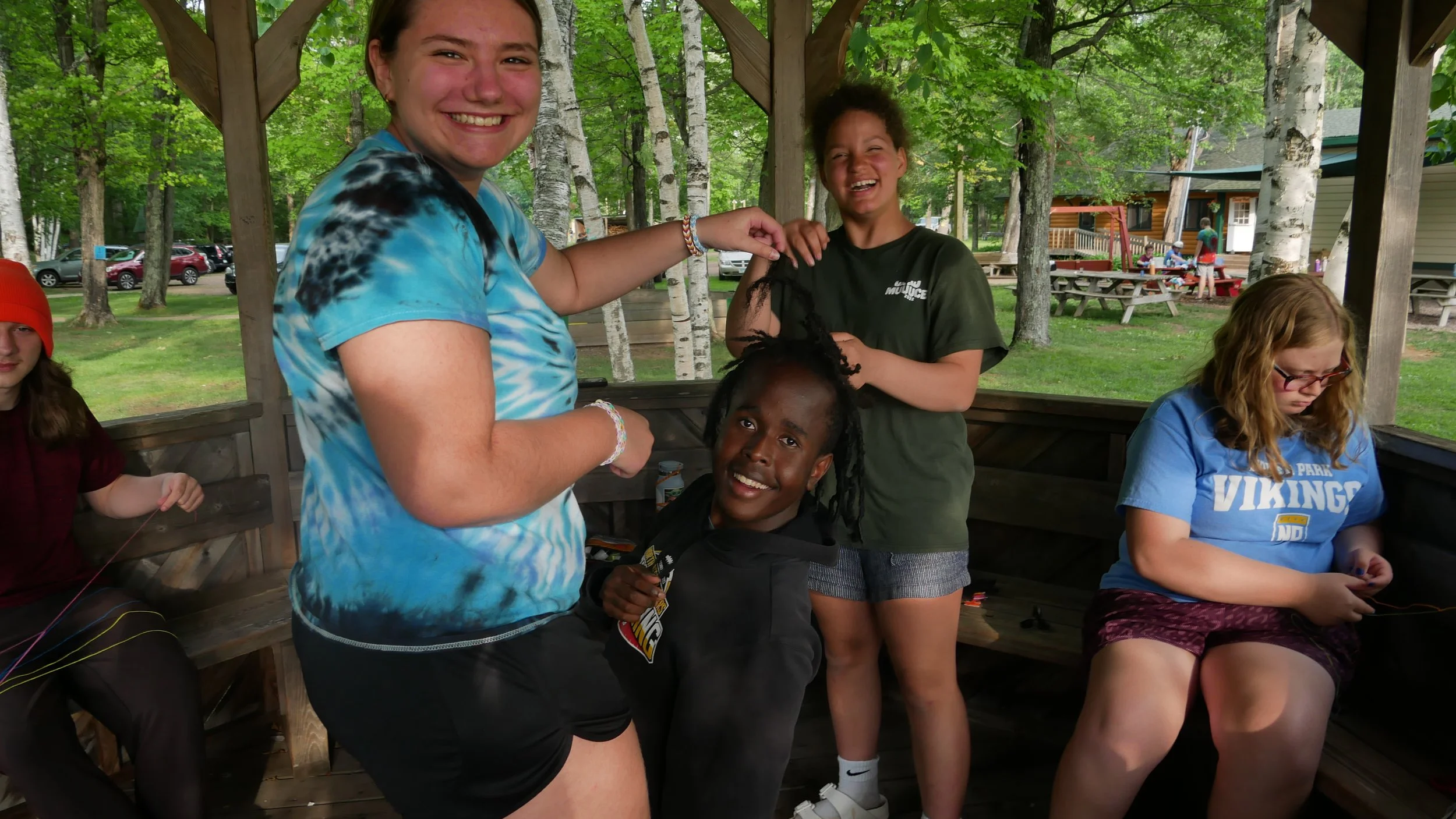Group of young teenagers sitting and standing in a wooden gazebo outdoors surrounded by trees. One girl in a blue tie-dye shirt is smiling and pointing at a boy with dreadlocks who is smiling, while another girl in a black t-shirt is laughing, and a girl in a blue Vikings shirt is looking at her phone.