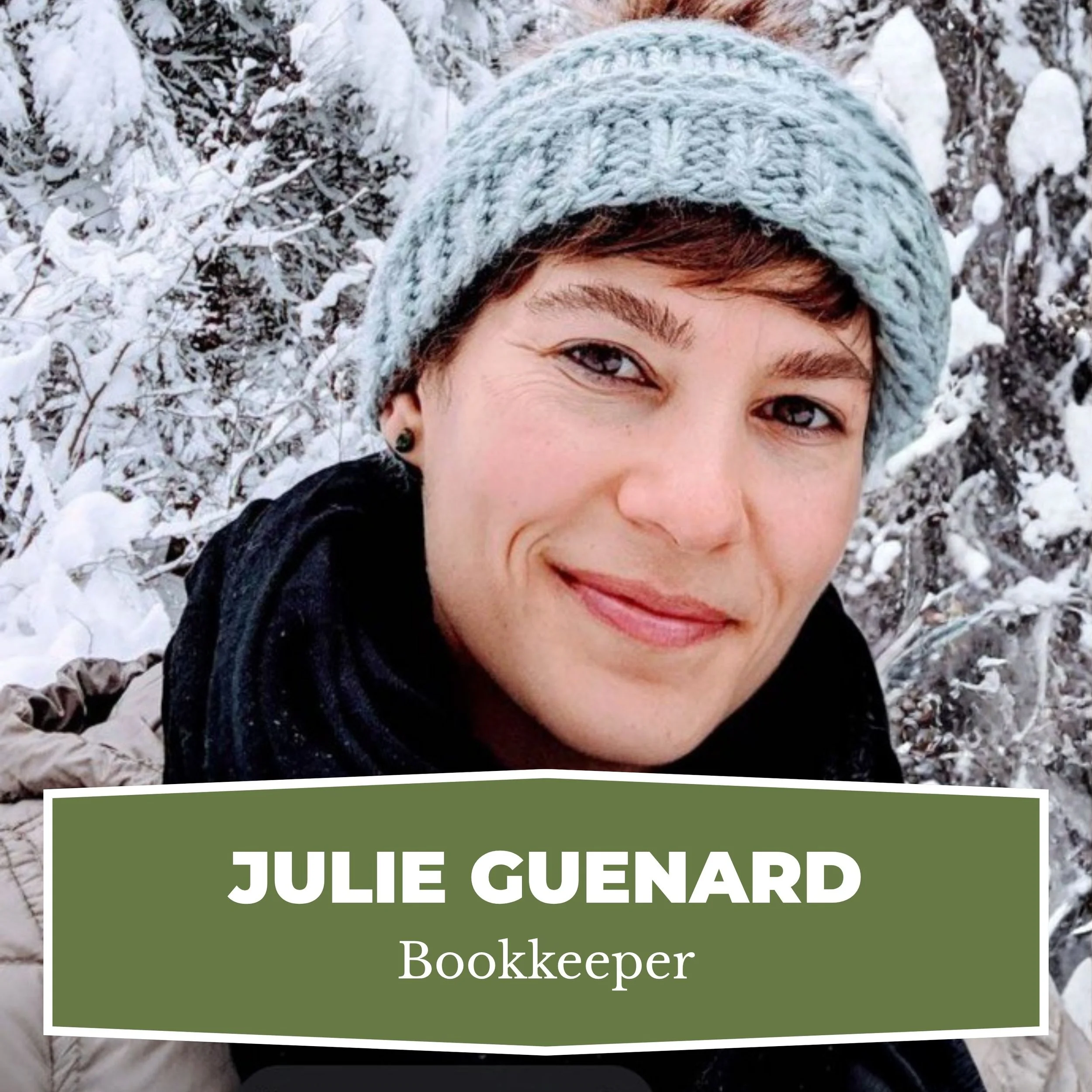 A woman with short brown hair and blue eyes wearing a knitted grey beanie, black scarf, and tan jacket, smiling in a snowy outdoor landscape.