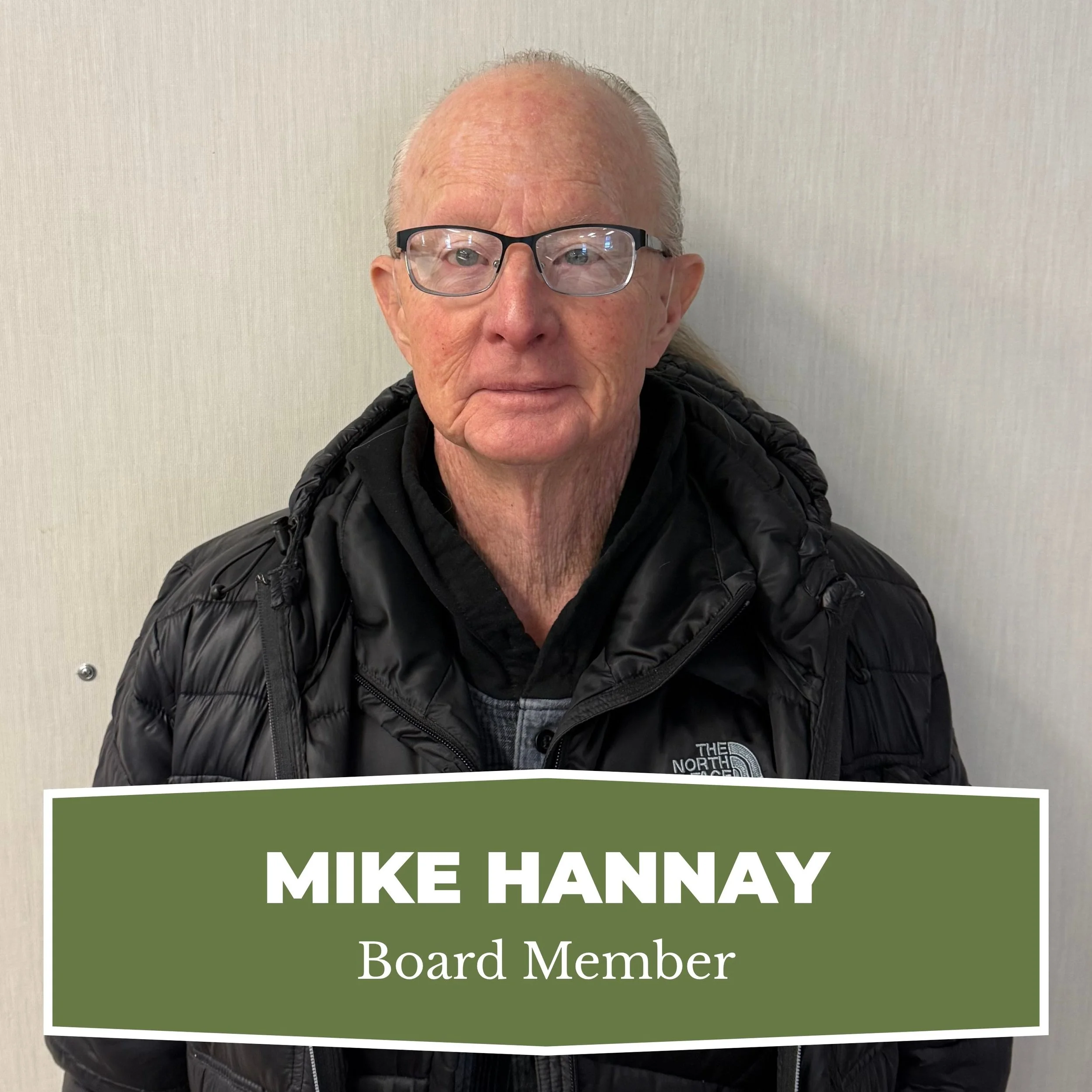 An elderly man with glasses wearing a black North Face jacket and a dark shirt, standing against a plain wall with a name tag that reads 'Mike Hannay, Board Member'.