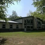 A large house with multiple windows and a deck, surrounded by trees and grass, on a sunny day.