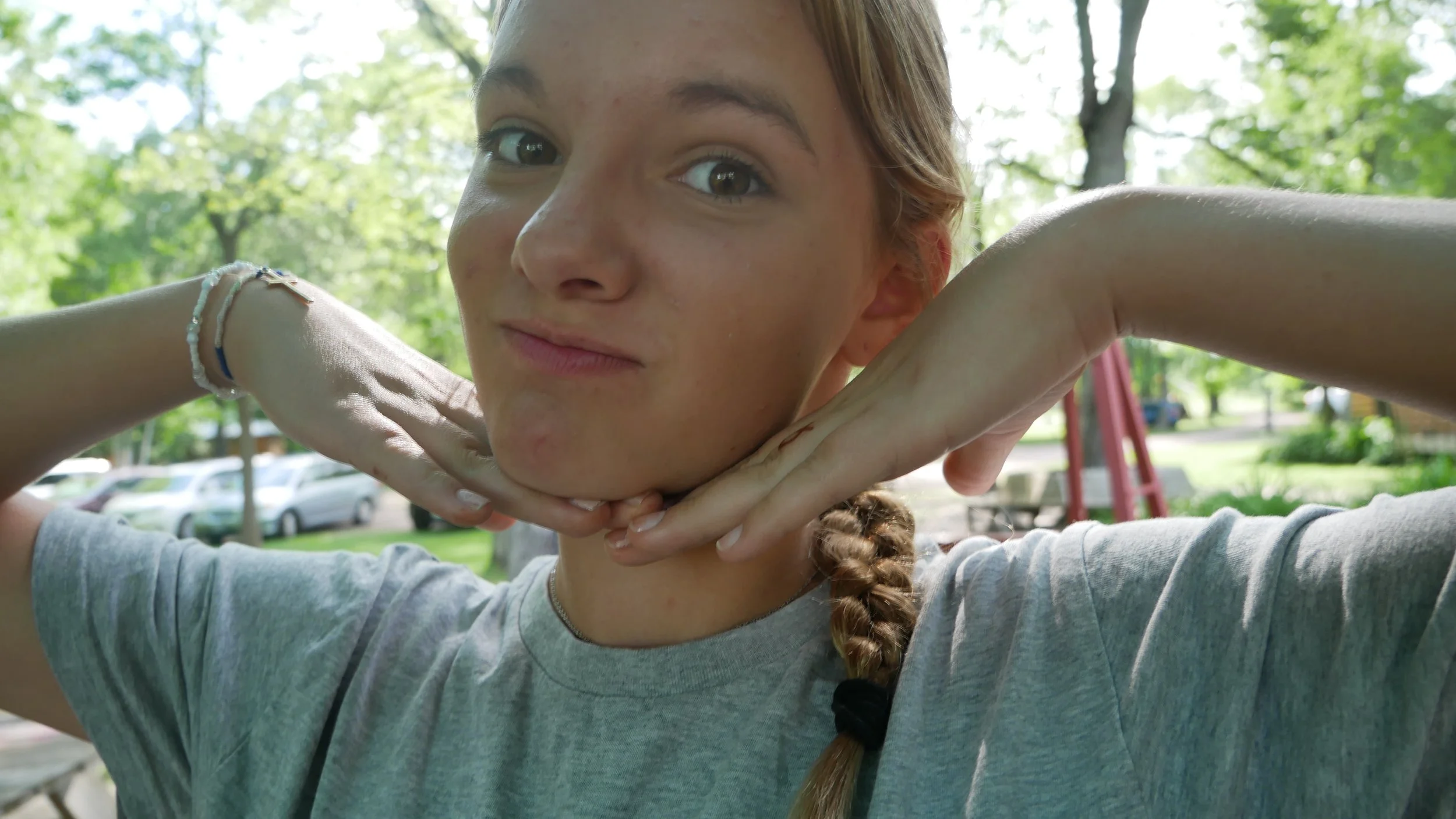 A young girl making a playful pose with her hands under her chin, smiling at the camera outdoors with trees and parked cars in the background.