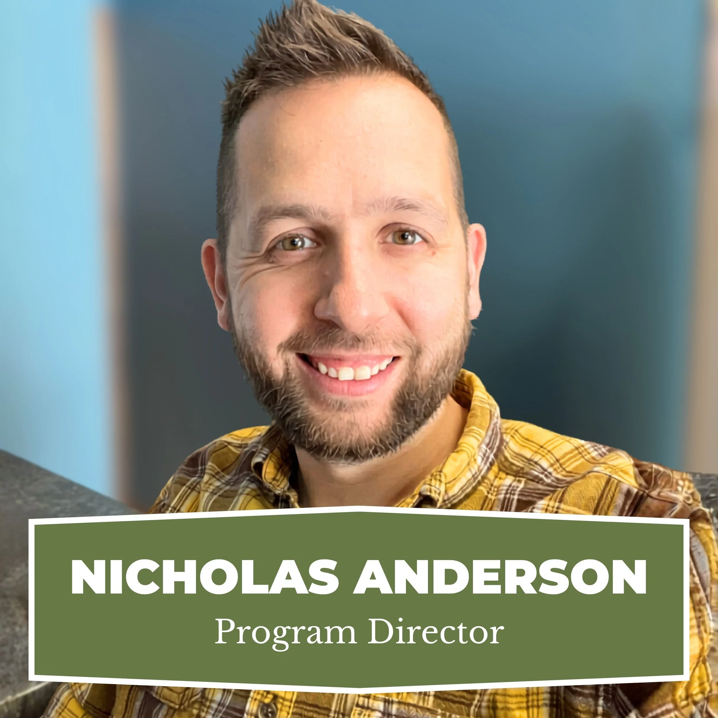 A smiling man with short brown hair and a beard, wearing a yellow and brown plaid shirt, with a name tag that reads 'Nicholas Anderson, Program Director'.