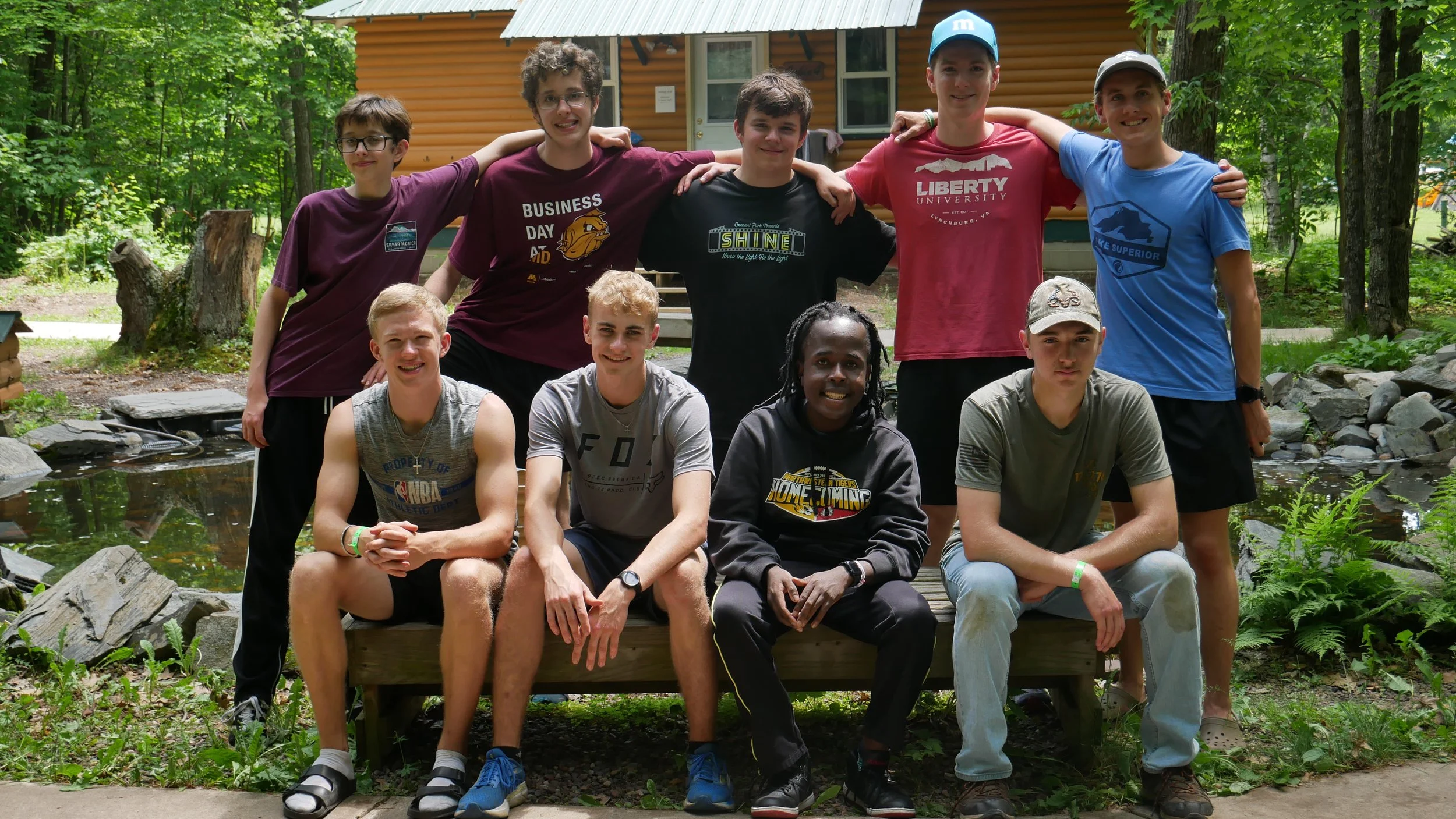 Group of ten teenage boys posing for a photo outdoors in a wooded area, some sitting on a wooden bench and others standing behind, with a small pond and a house in the background.
