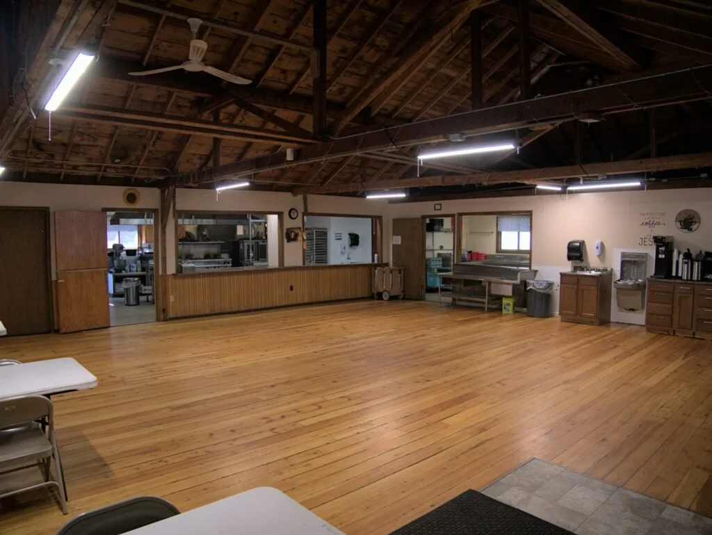 Large, open community room with a wooden floor and exposed wooden ceiling. There are tables and chairs in the foreground, and a serving area with a water fountain, coffee station, and kitchen counters along the back wall. Fluorescent lights illuminate the room.