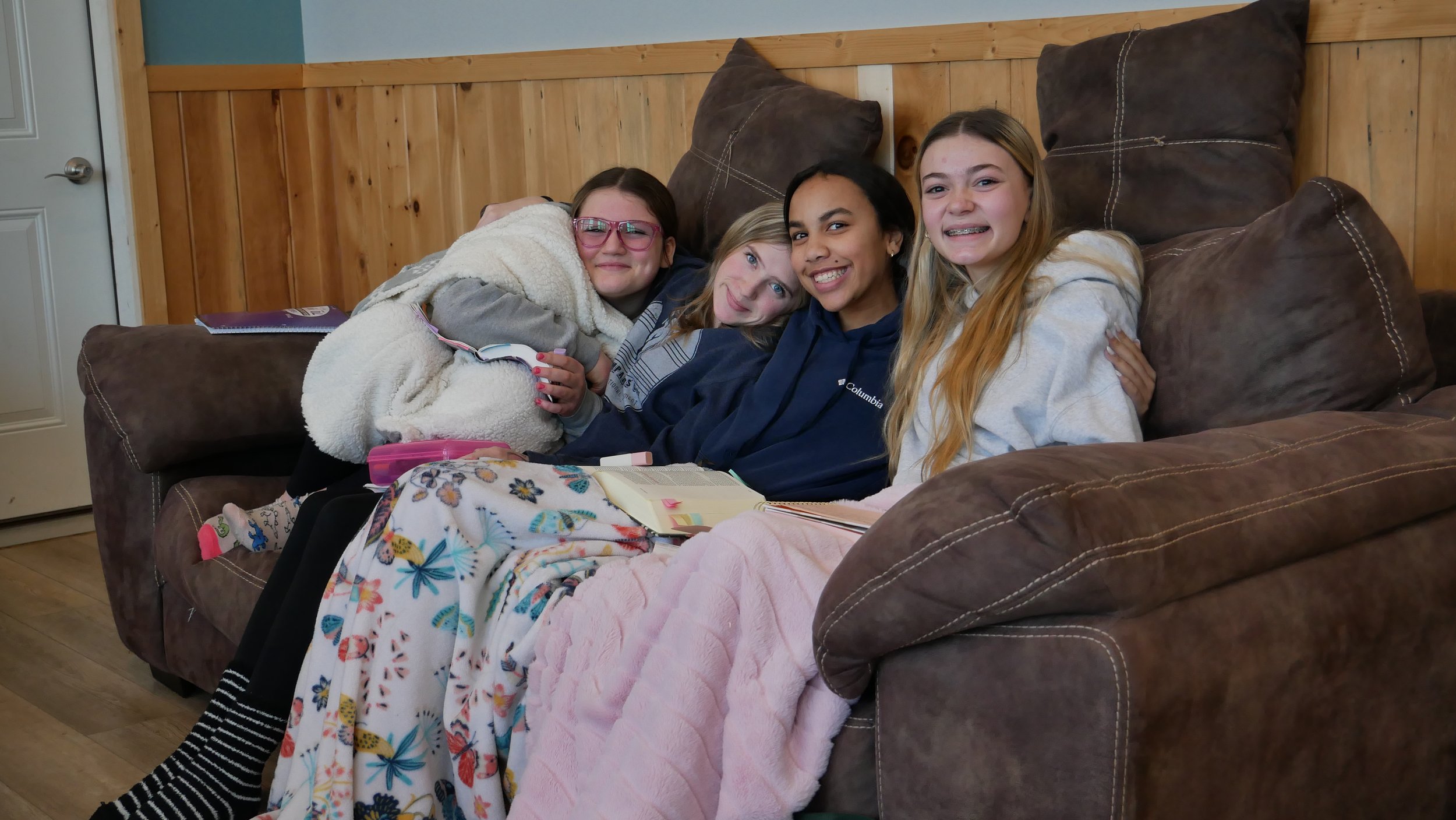 Four girls sitting on a brown leather couch in a cozy room with wooden paneled walls, smiling at the camera.