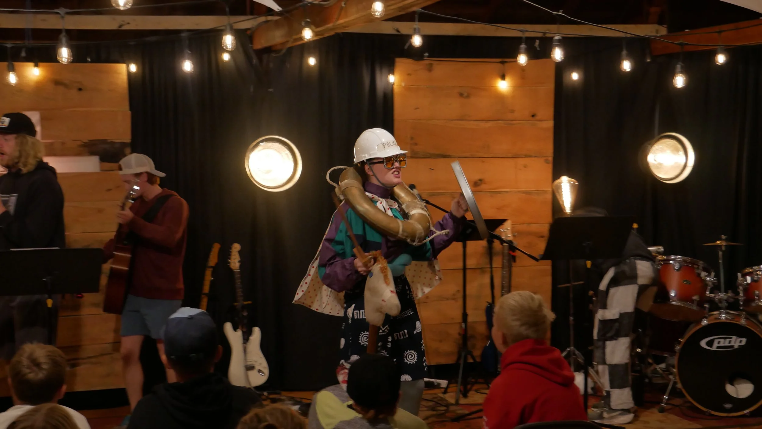 A band performing on stage with a woman dressed in an eclectic costume resembling a space explorer, wearing a white helmet labeled 'POLICE', sunglasses, a colorful jacket, and carrying a small purse. The stage has wooden wall paneling, string lights, guitars, and a drum set, with audience members seated in front.