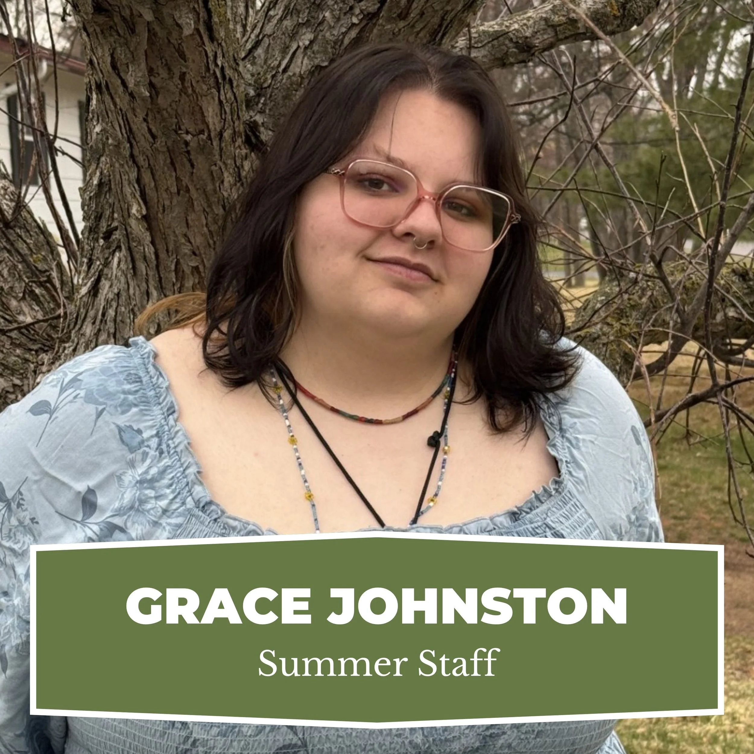 A young woman with dark hair, light skin, and glasses, standing outdoors near a tree with bare branches, wearing a light blue dress and layered necklaces, smiling slightly, with a sign that reads "Grace Johnston, Summer Staff" in front of her.