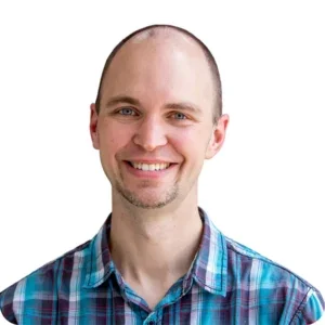 A smiling man with a shaved head wearing a blue plaid shirt, facing forward against a plain white background.