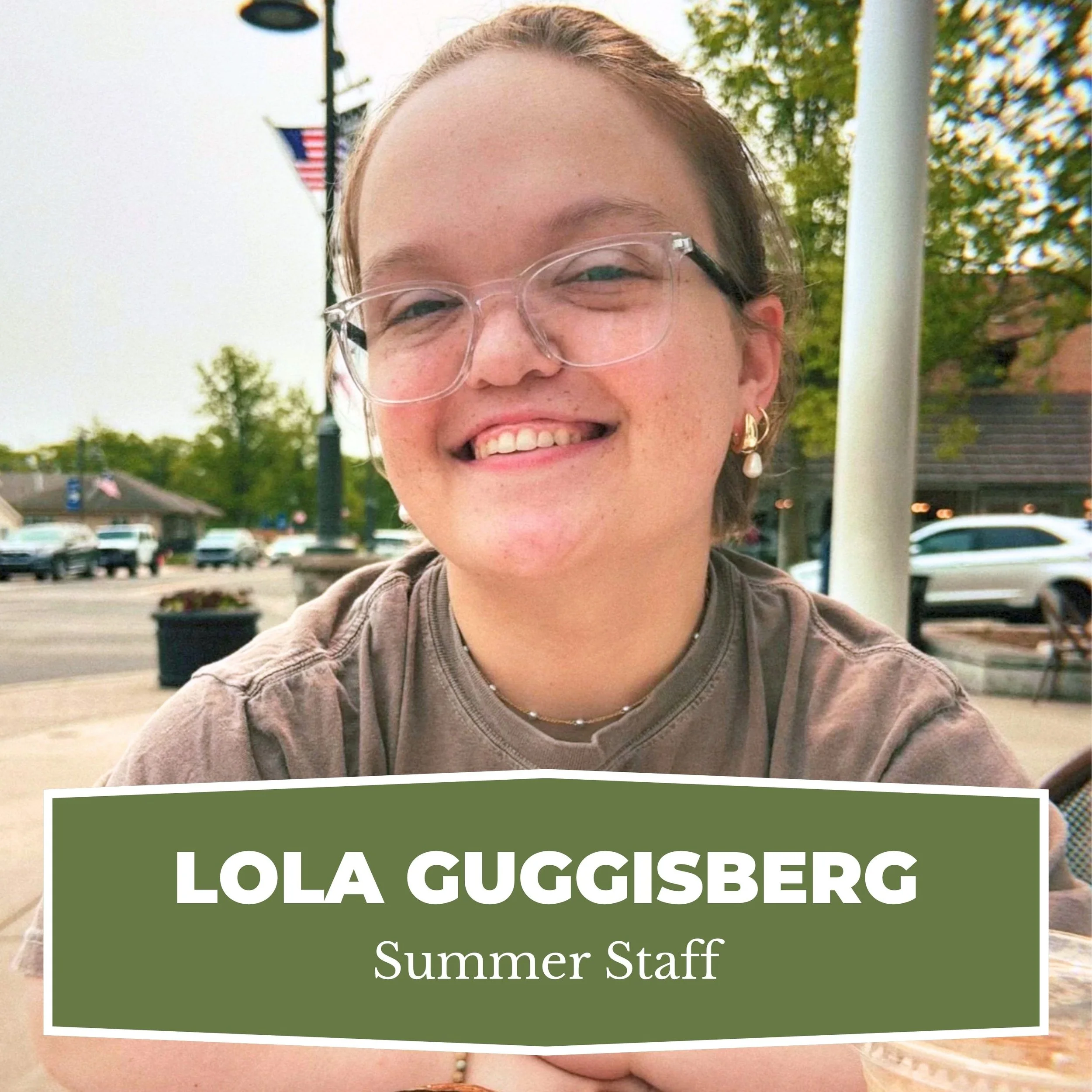 Smiling young woman with glasses sitting at an outdoor table, wearing earrings and a brown shirt, with a sign reading "Lola Guggisberg, Summer Staff." There are cars and trees in the background.