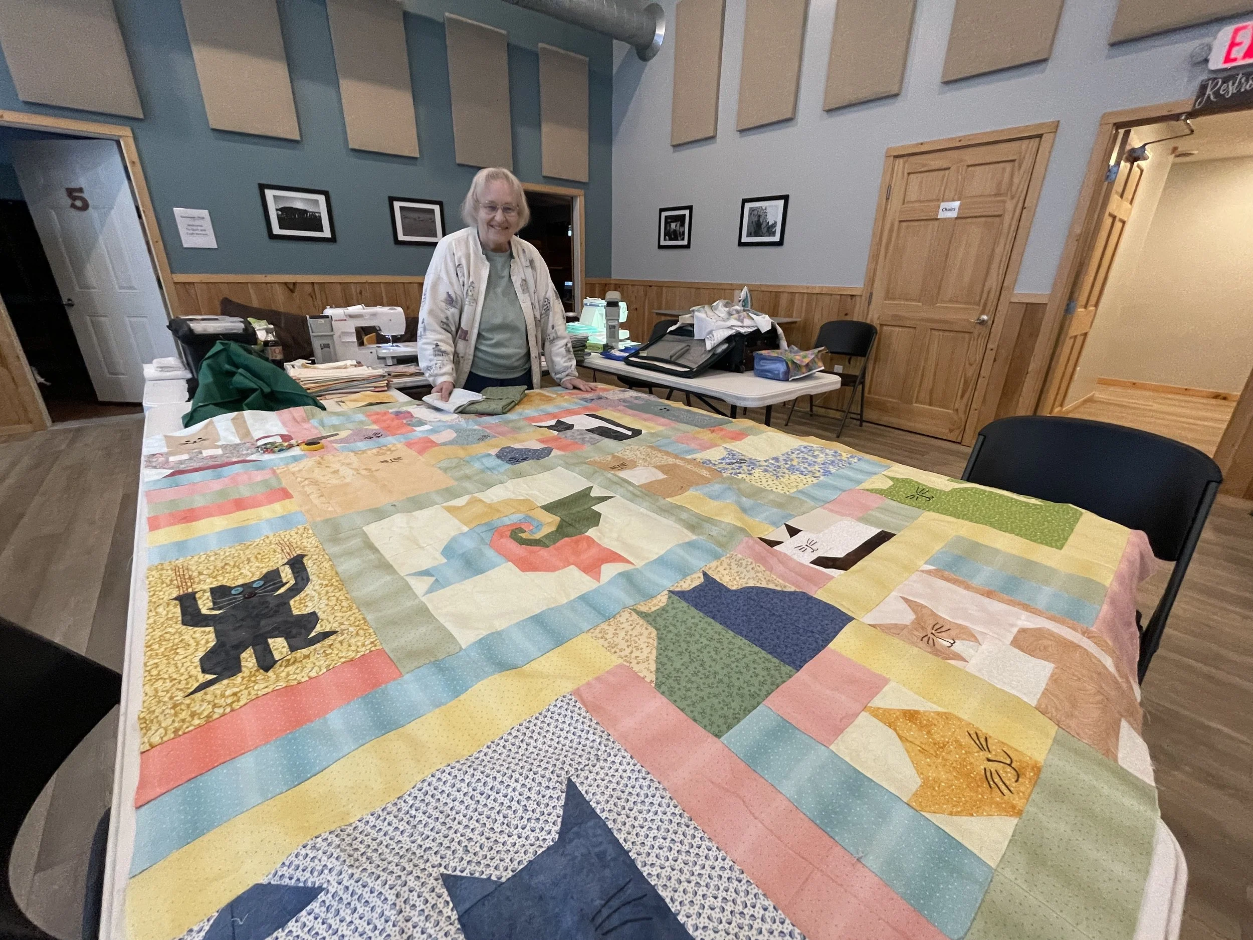 A woman standing behind a large colorful quilt on a table in a room with wood-paneled walls and framed black and white photographs.