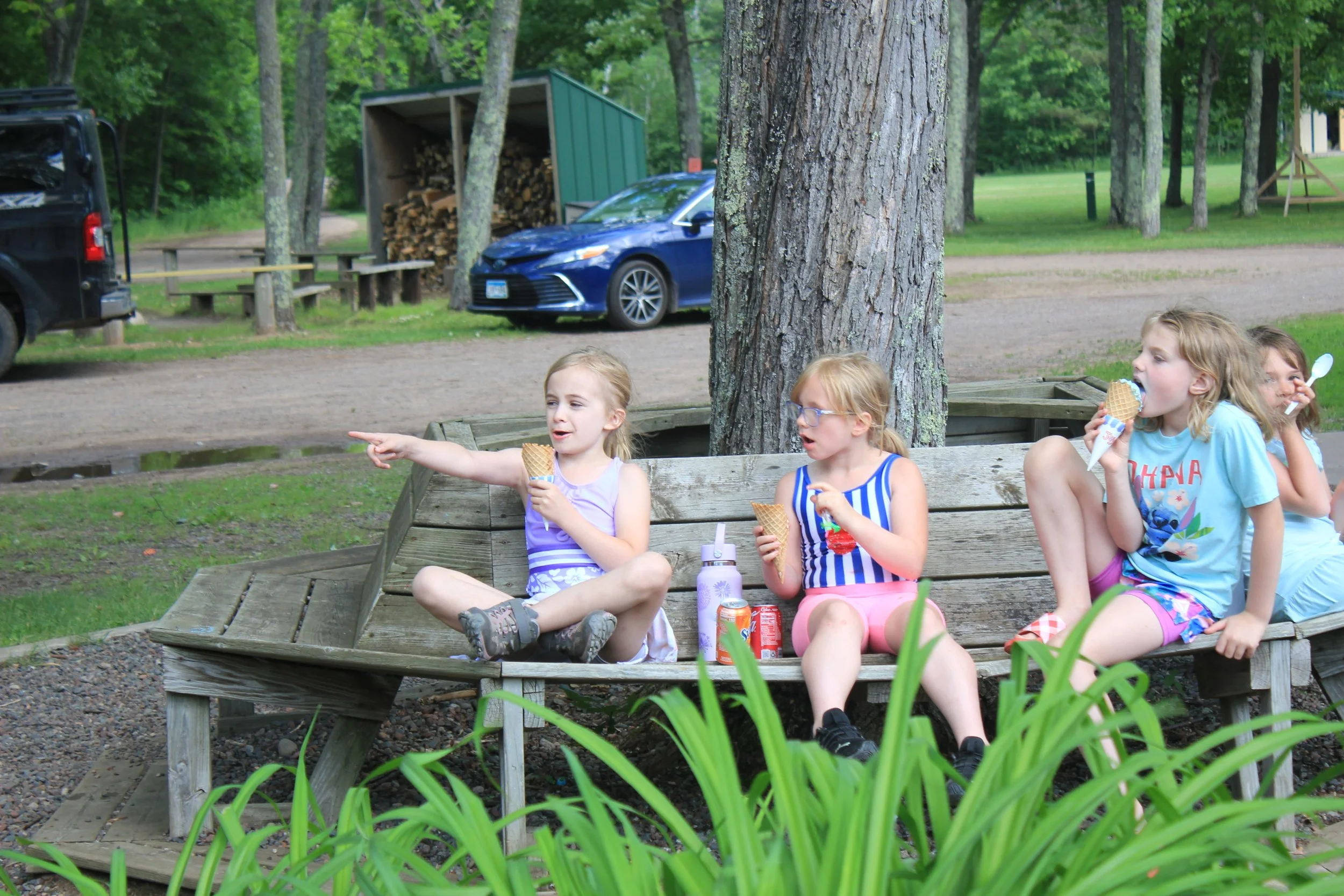 Four young girls sitting on a wooden bench outdoors, eating ice cream cones, with trees and parked cars in the background.