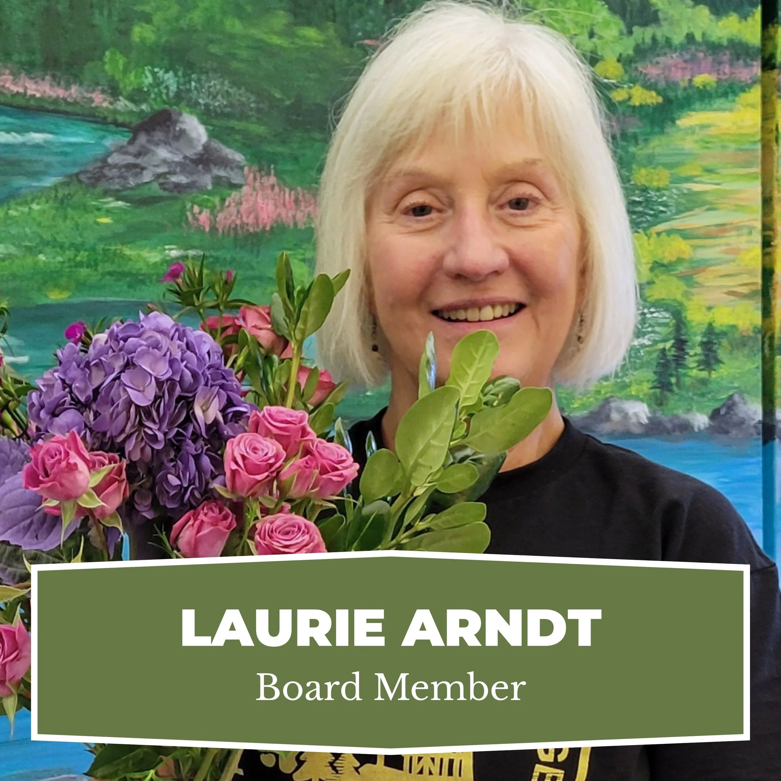 A woman with short white hair smiling and holding a bouquet of pink roses and purple hydrangeas, standing in front of a colorful landscape painting. A name badge or label with her name 'Laurie Arndt' and title 'Board Member' is visible.