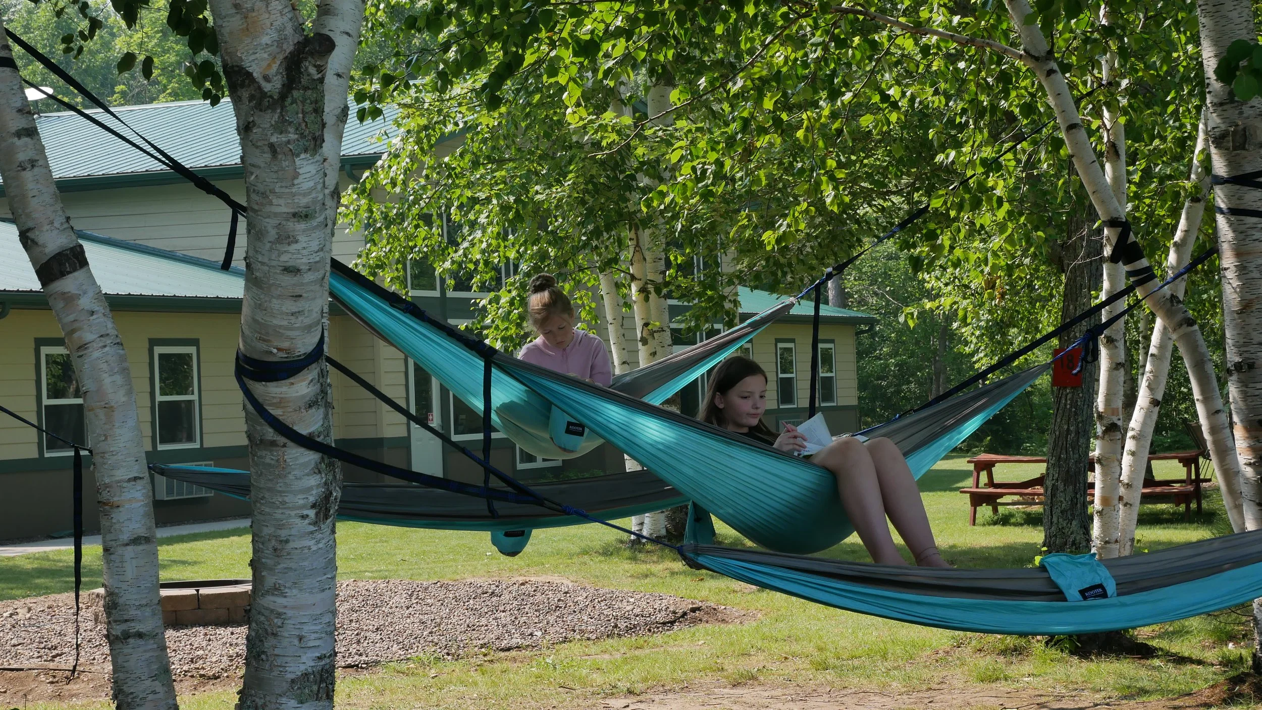 Two girls relaxing in separate hammocks hung between trees in a backyard with green grass, trees, and a yellow house in the background.