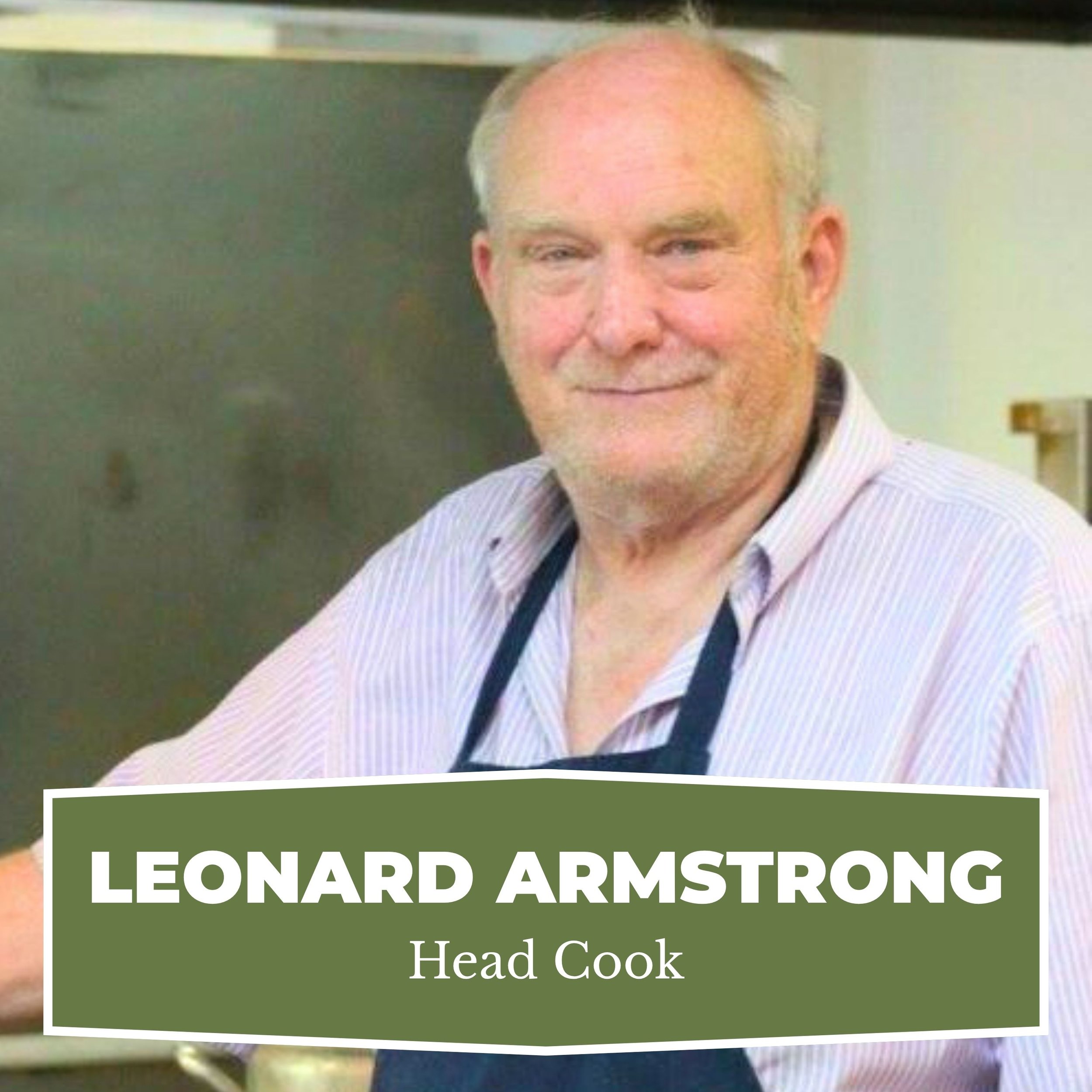 A senior man with short gray hair, beard, and mustache, wearing a striped button-up shirt and a dark apron, smiling in a kitchen setting.