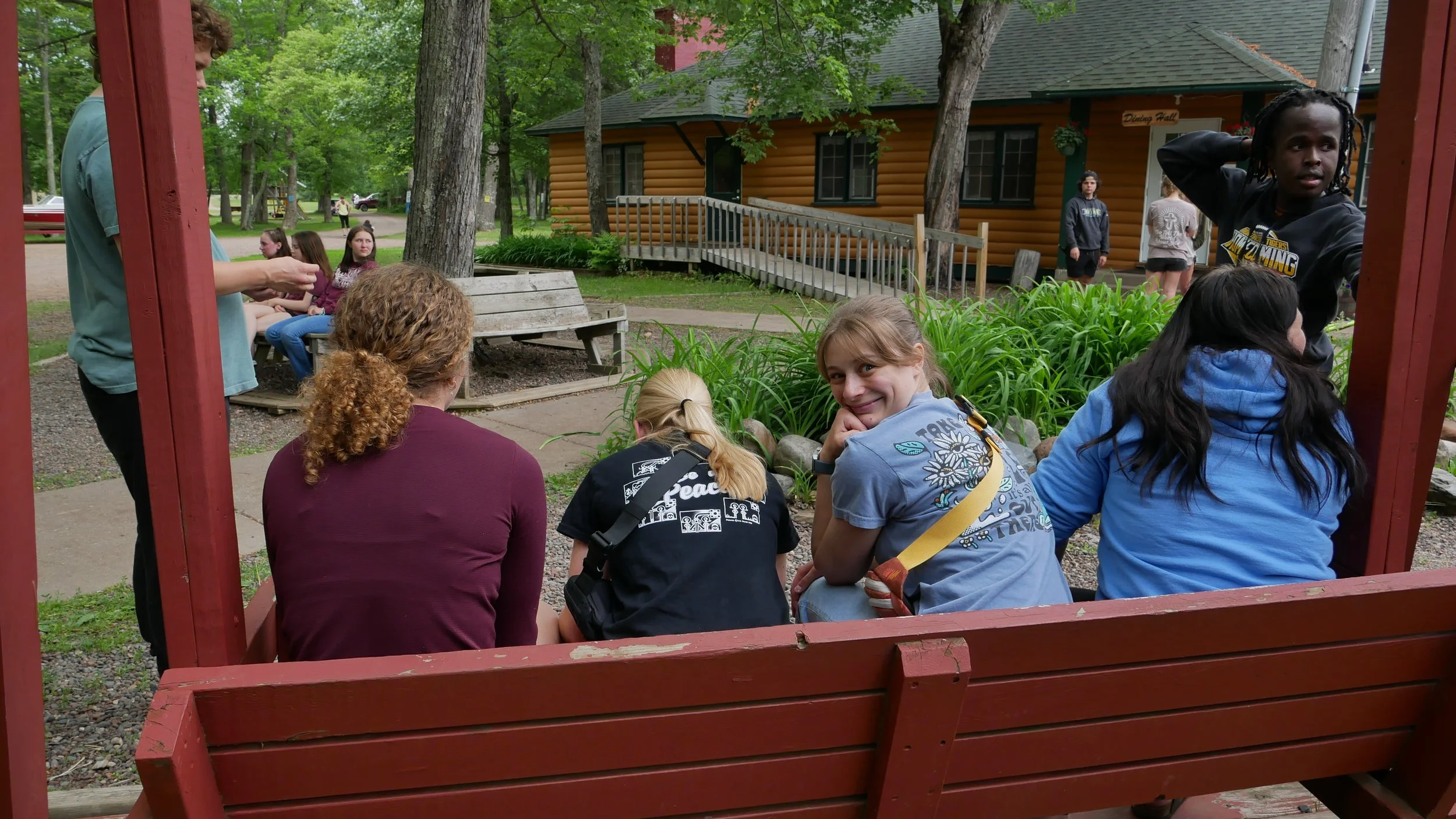 A group of children and teenagers sitting on a wooden bench in a park, near a garden with green plants, with some standing and talking in the background, surrounded by trees and a log cabin style building.
