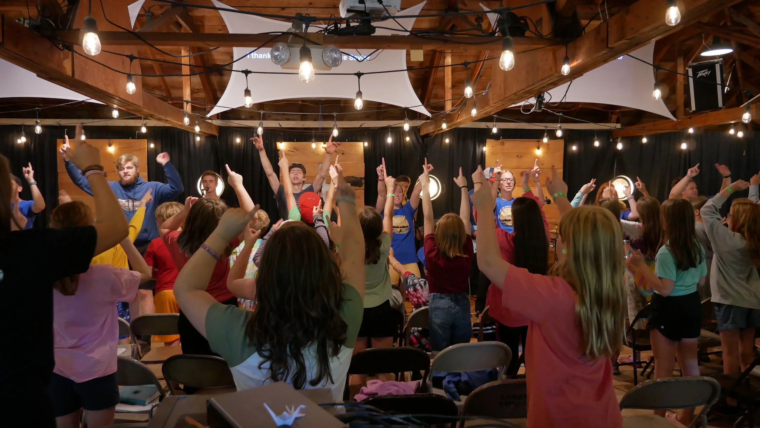 A group of people, mostly children and teenagers, dancing and raising their hands in the air at an indoor event with wooden walls and hanging string lights.